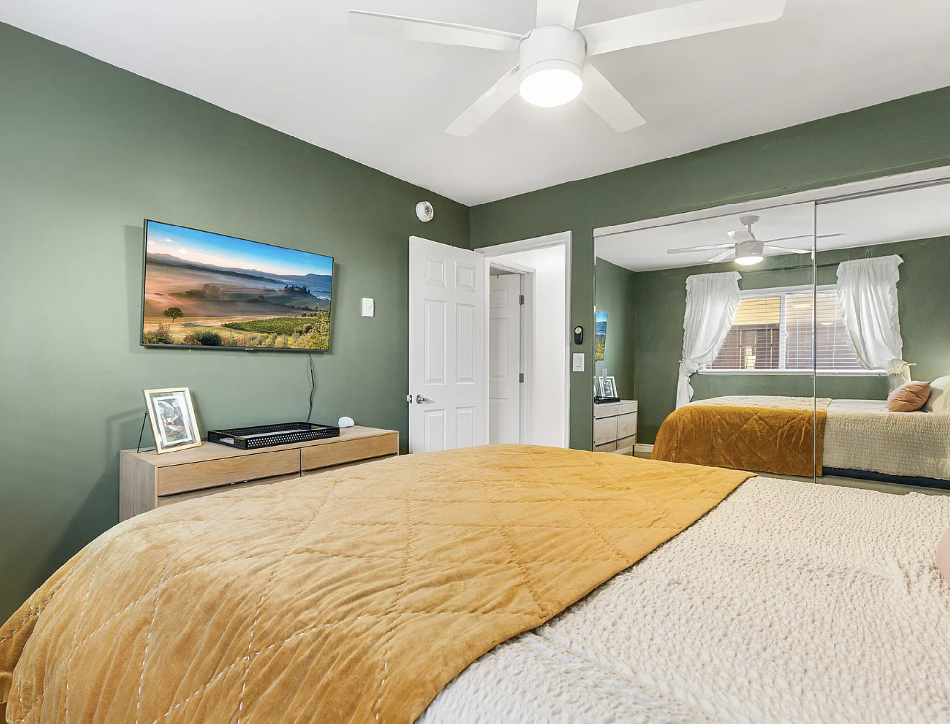 Bedroom with green walls, mounted TV, mirrored closet, dresser, white ceiling fan, and gold bedspread.
