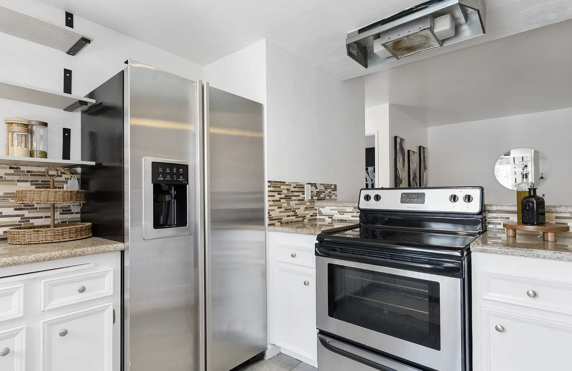 Modern kitchen with stainless steel refrigerator and oven, granite countertops, white cabinets, tile backsplash, and open shelves with jars and baskets.