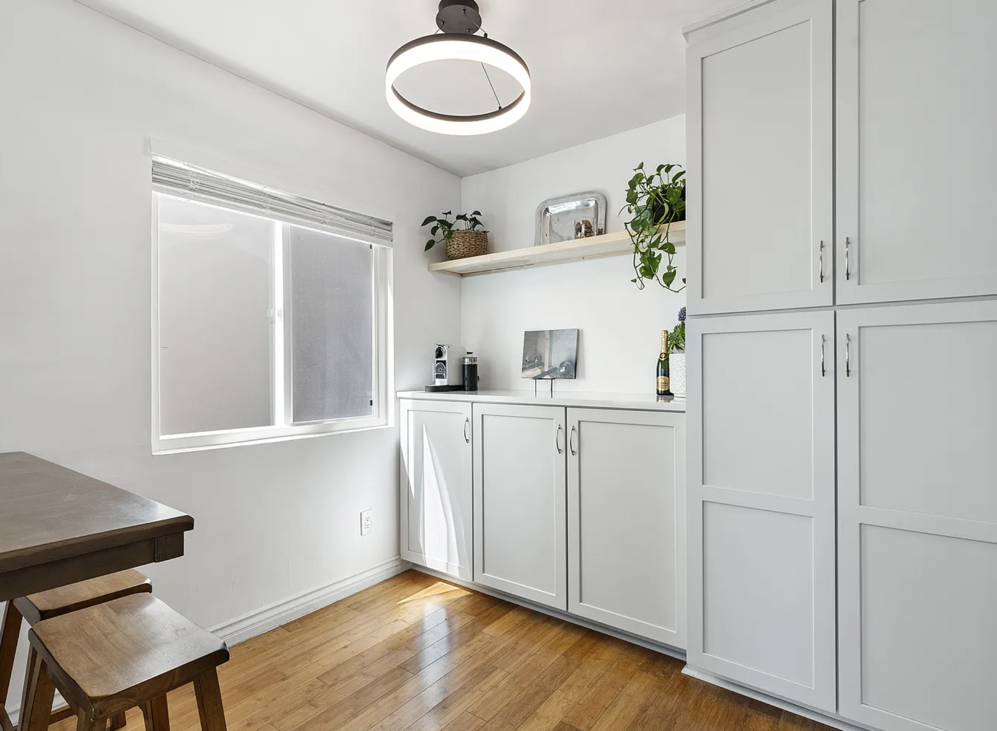 Minimalist kitchen with white cabinets and countertop, bamboo floor, window, modern circular light fixture, wooden table and stools, potted plants and coffee maker, and decorative items on shelf and countertop.