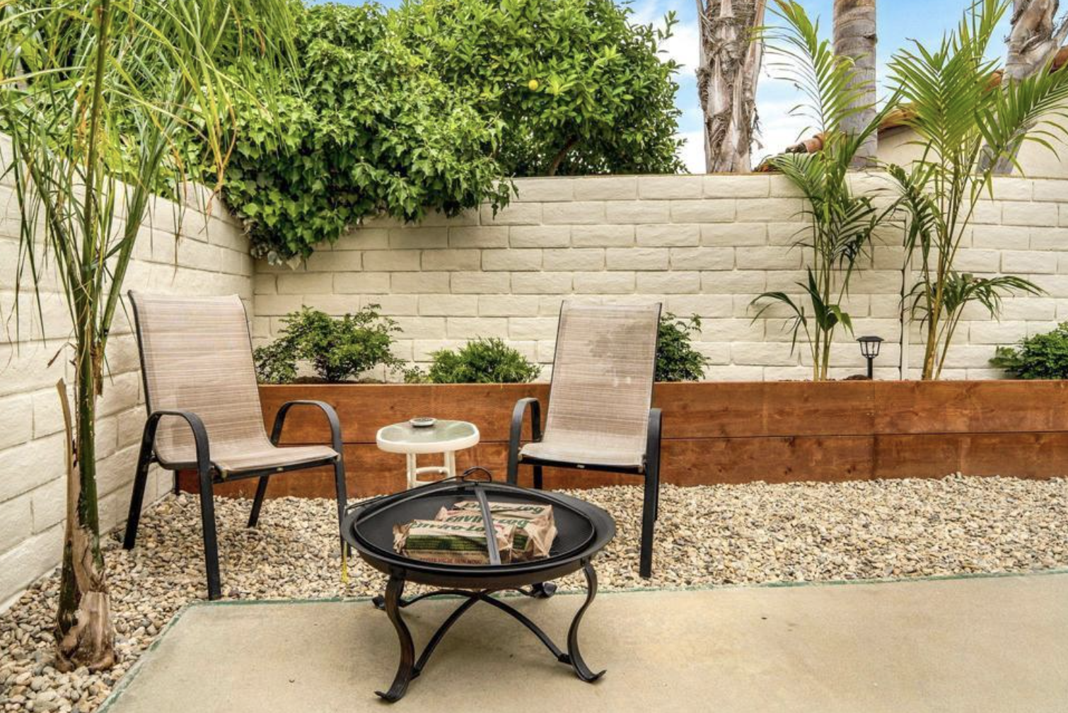 Outdoor patio with two chairs, a small table, and a fire pit on pebble-strewn ground, surrounded by a white brick wall and greenery.