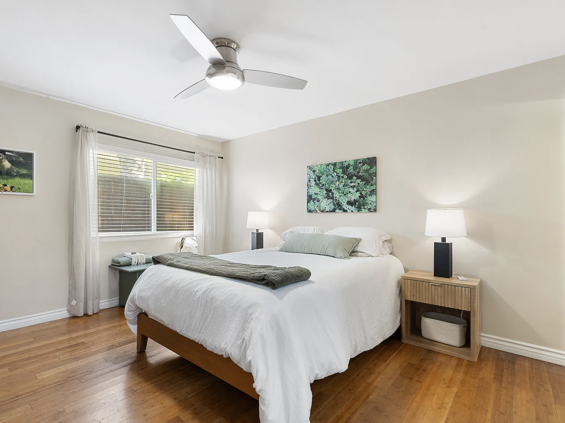 Modern bedroom with wooden floor, white bedding, ceiling fan, and bedside lamps. Features artwork and window with sheer curtains.