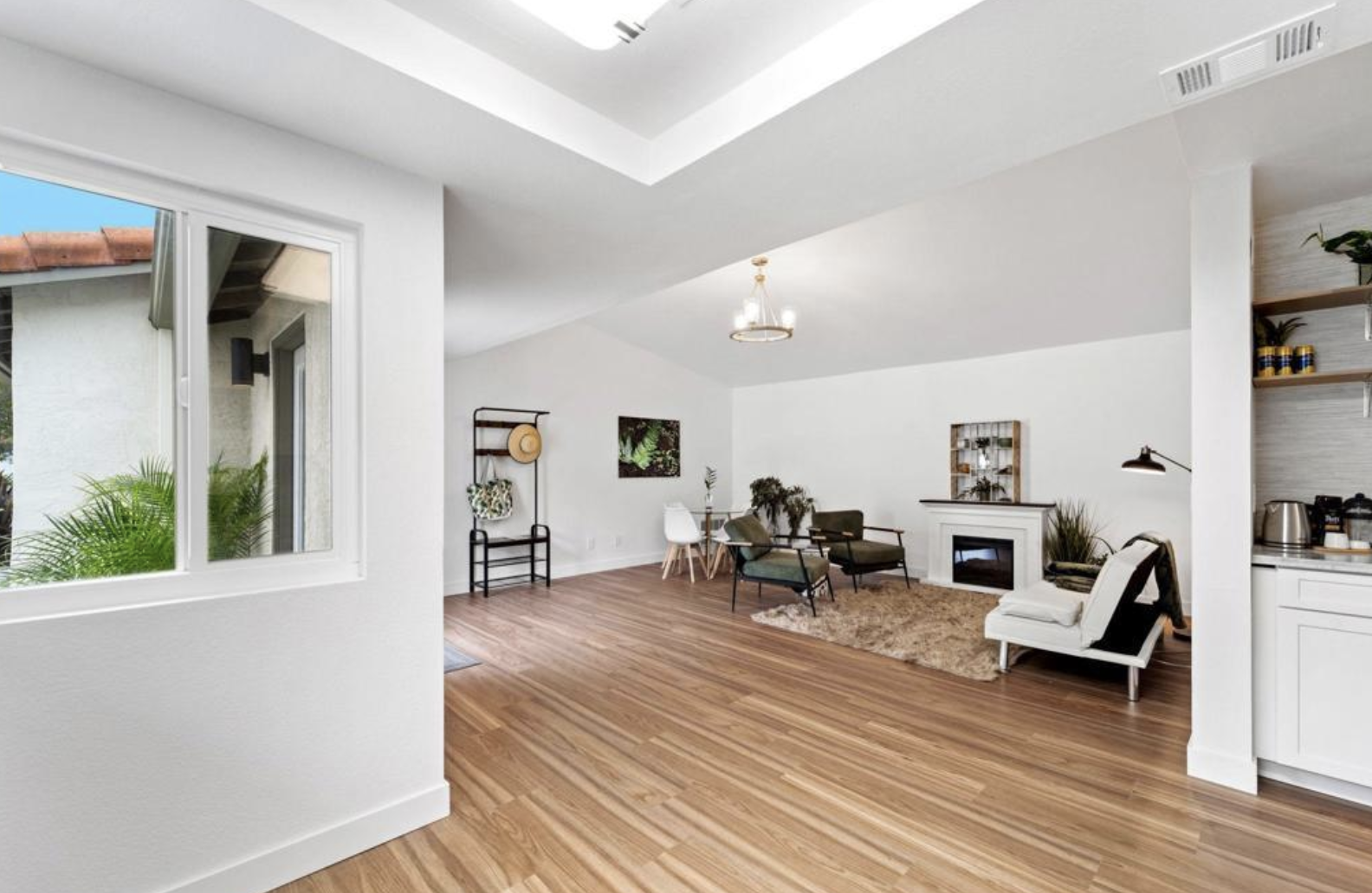 Modern living room with wood flooring, white walls, a window with plants outside, and a cozy seating area including a fireplace, armchairs, and a small rug. There is a hat and bag on a rack near the entrance.