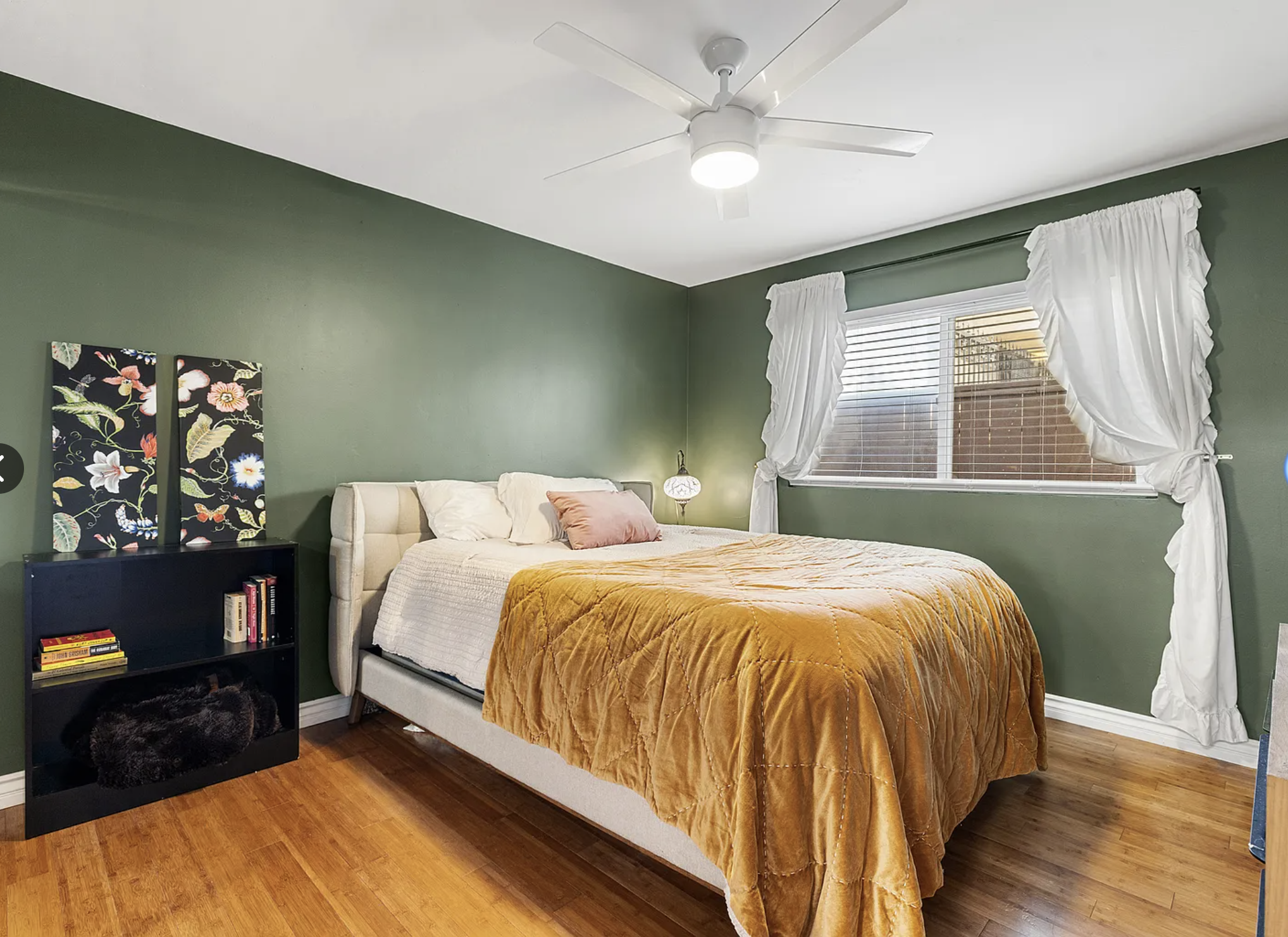 Bedroom with green walls, bed with mustard yellow comforter, black shelf with books and floral art, white ceiling fan, window with white curtains.