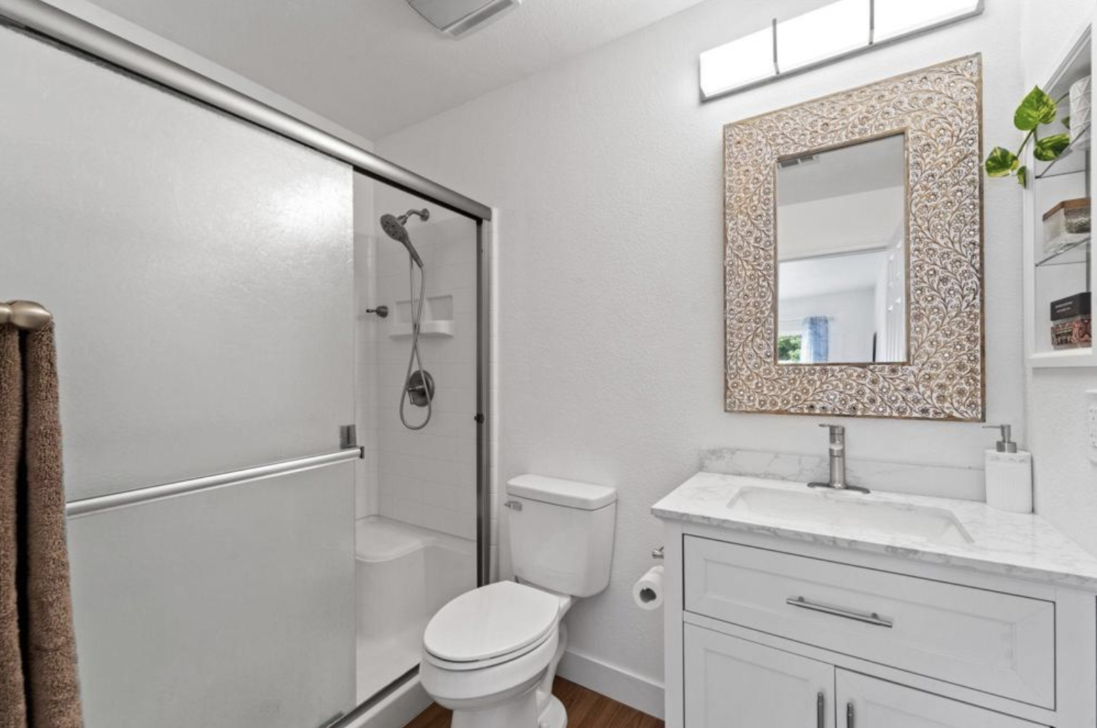 Modern bathroom with a glass shower, white toilet, marble countertop sink, ornate mirror, and wall shelves with toiletries.