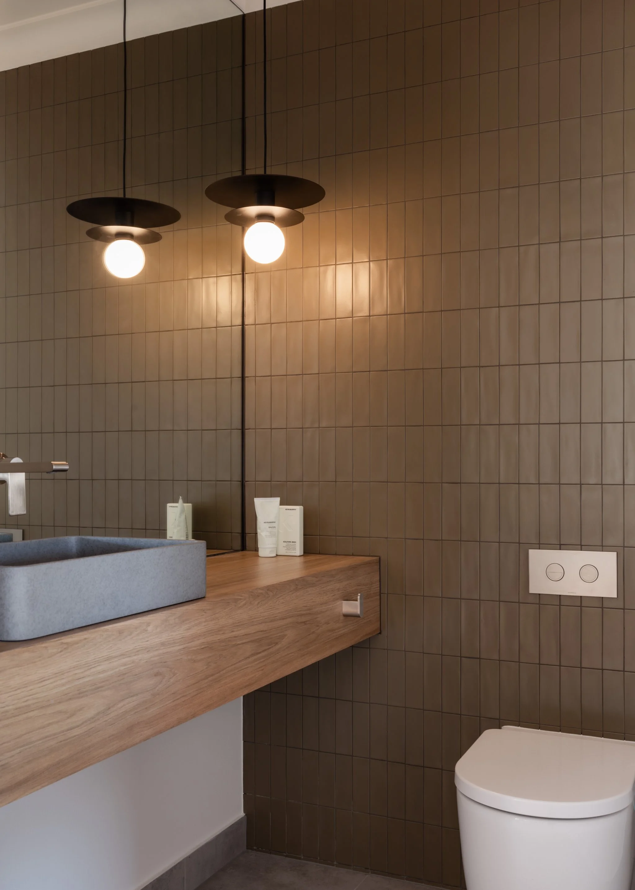 Modern bathroom with brown tiled wall, wooden countertop, ceramic sink, and white toilet. Two black pendant lights hang from the ceiling, illuminating the space.