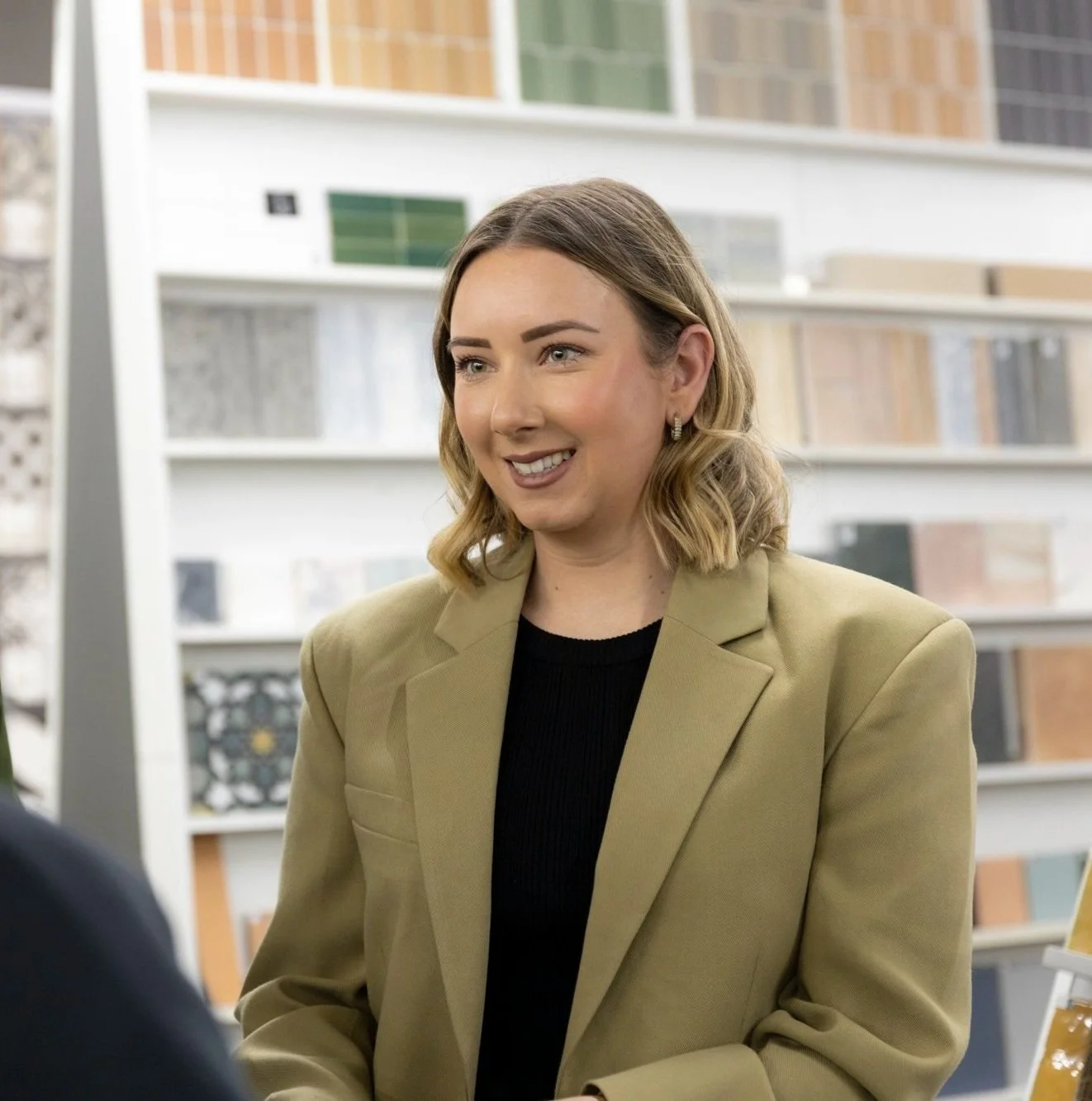 A woman with shoulder-length blonde hair and light skin, wearing a beige blazer over a black top, smiling in a store with shelves of tile samples in the background.