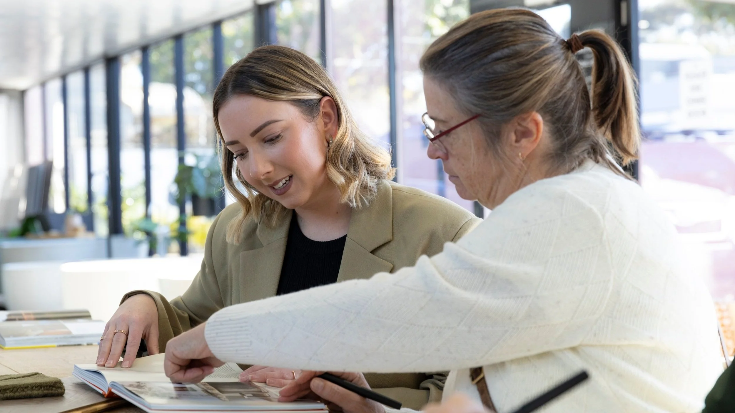 Two women sitting at a table, looking at a book and discussing. The woman on the left is younger with shoulder-length blonde hair, wearing a beige blazer. The woman on the right is older, with glasses and her hair in a ponytail, wearing a white sweater.