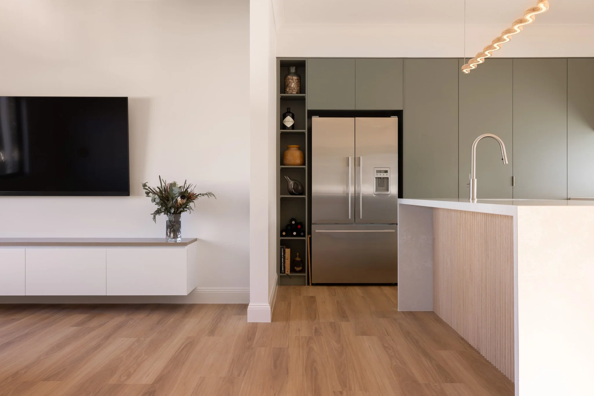 Modern kitchen and living room with wood flooring, a flat-screen TV on the wall, a floral arrangement on a white console, and a stainless steel refrigerator with built-in shelves