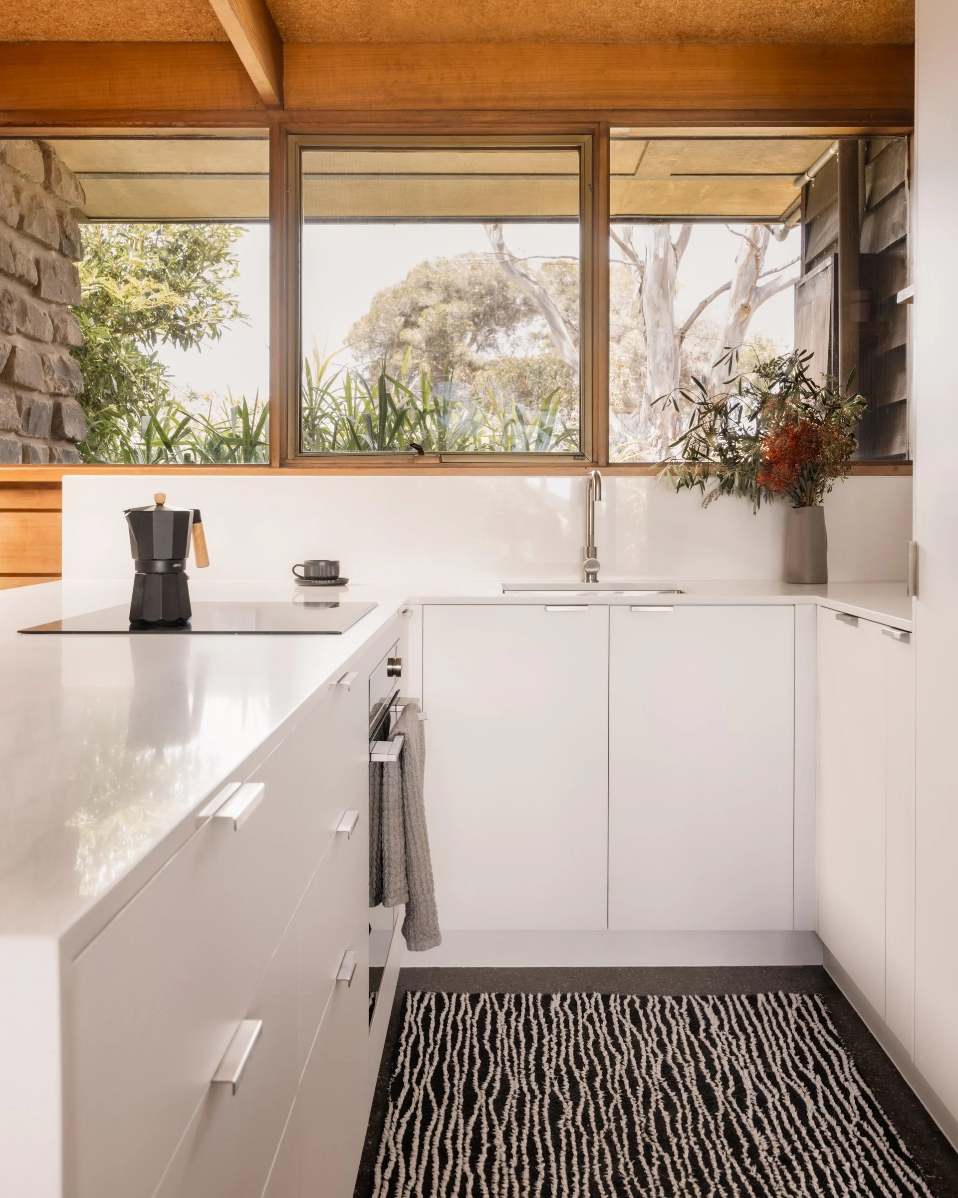 A modern kitchen with white cabinets, a bowl of coffee, a moka pot on the countertop, large windows showing trees outside, a gray and black striped rug, and a flower vase with plants.