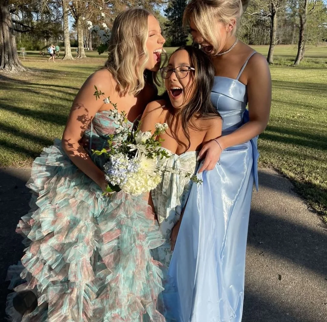 Three women dressed in formal gowns are standing together outdoors, smiling and laughing. One holds a bouquet of white and blue flowers.