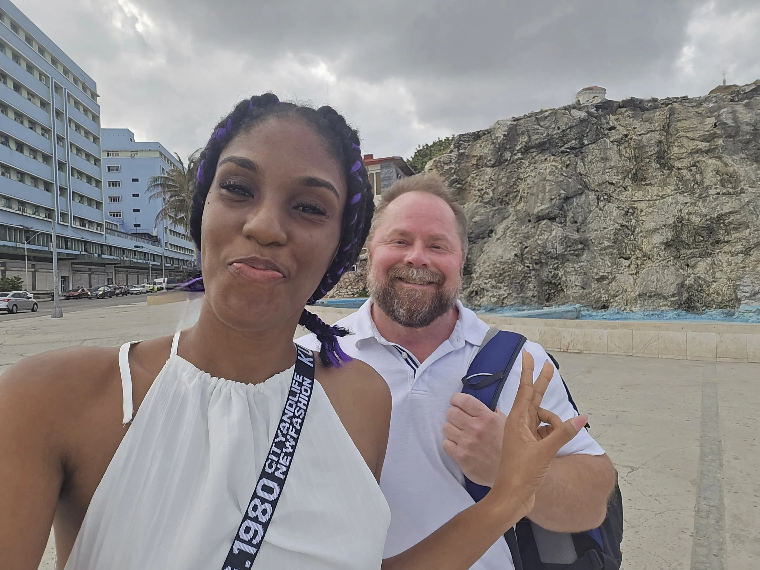 Two people posing for a selfie outdoors with buildings and a rocky formation in the background.
