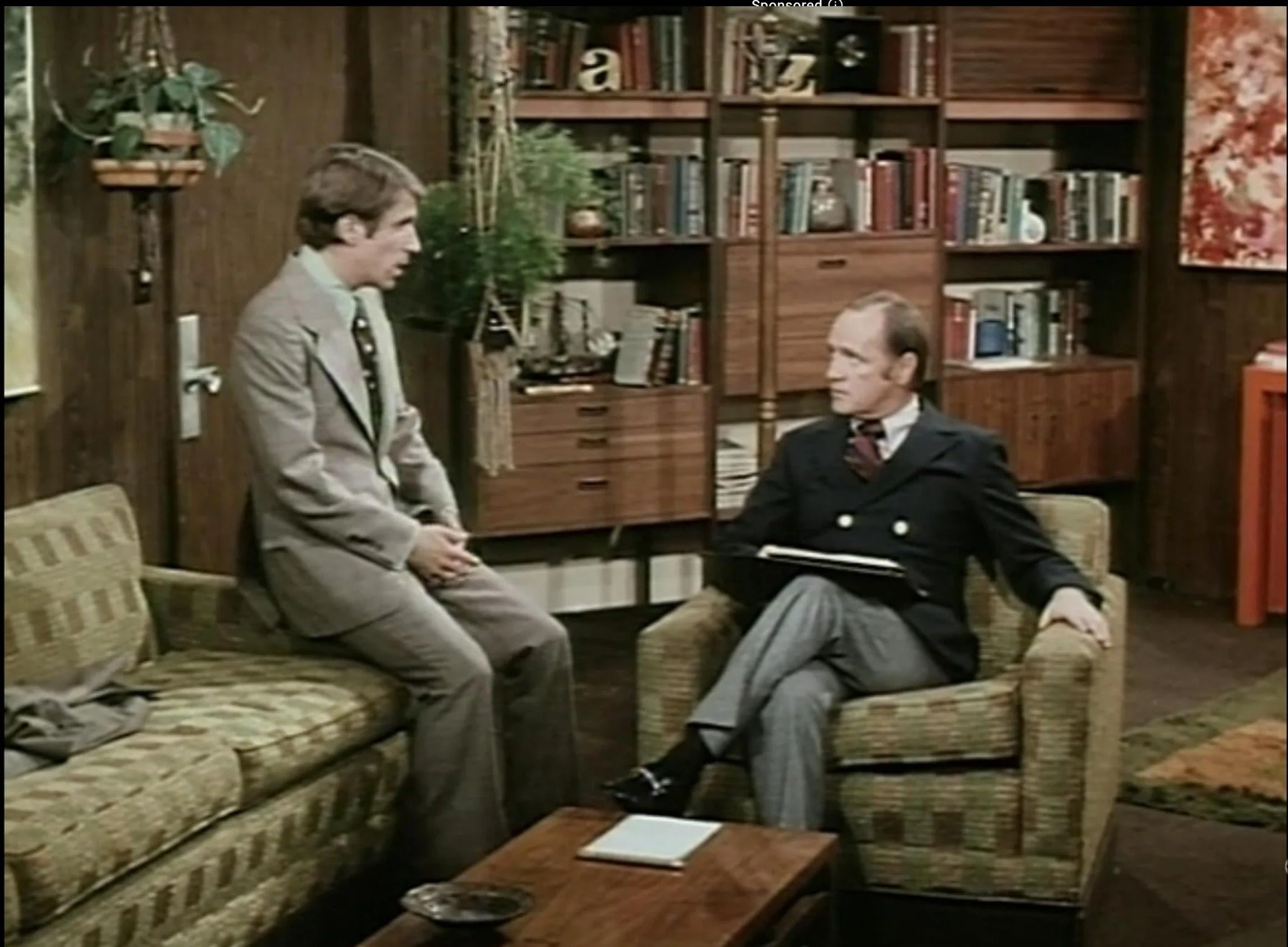 Two men in a vintage office with bookshelves, one seated, one leaning against a couch, wearing suits.