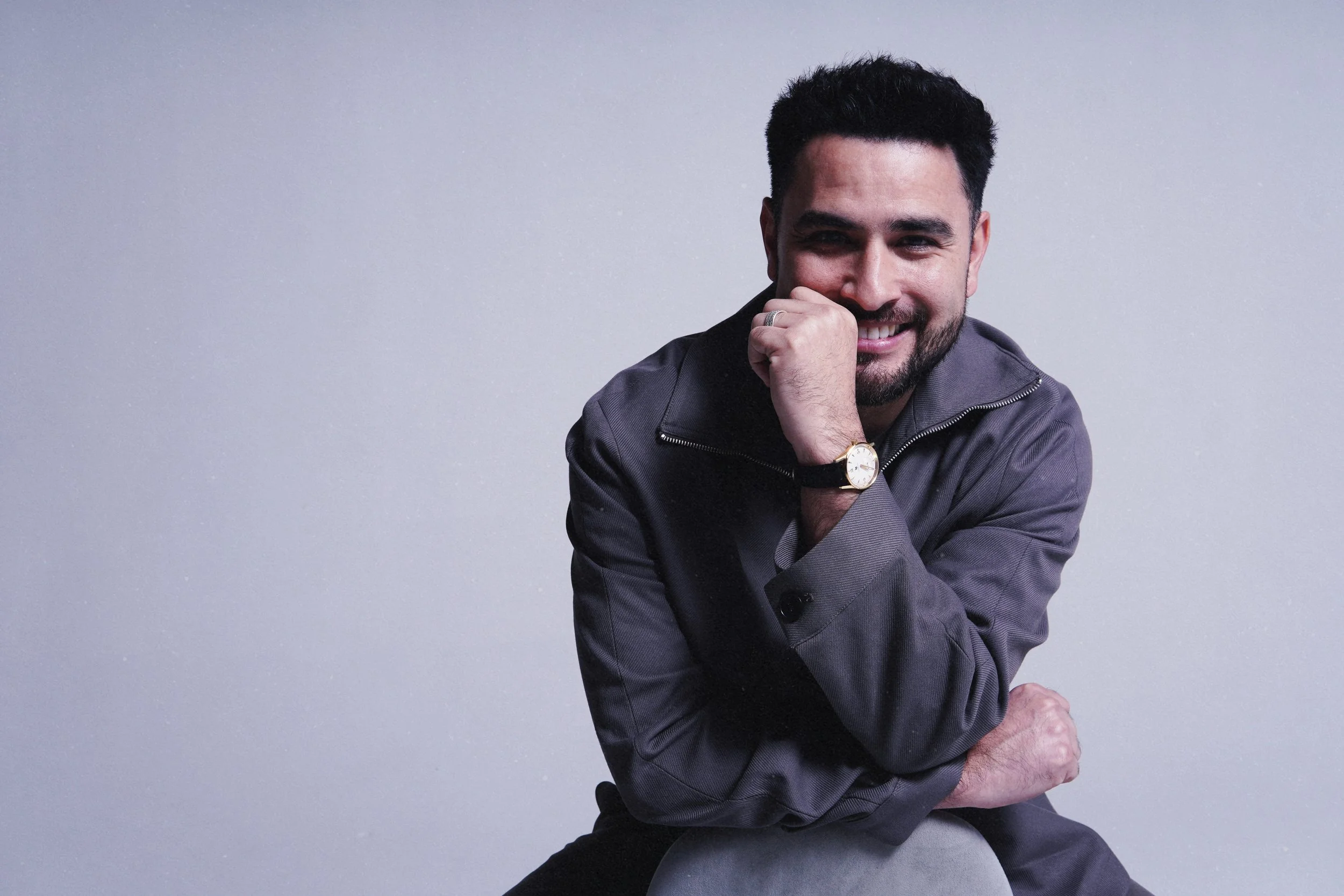 A man with dark hair and a beard smiling, wearing a gray jacket, a white shirt, a gold watch, and a ring, posing with one arm flexed and hand near his face against a plain gray background.
