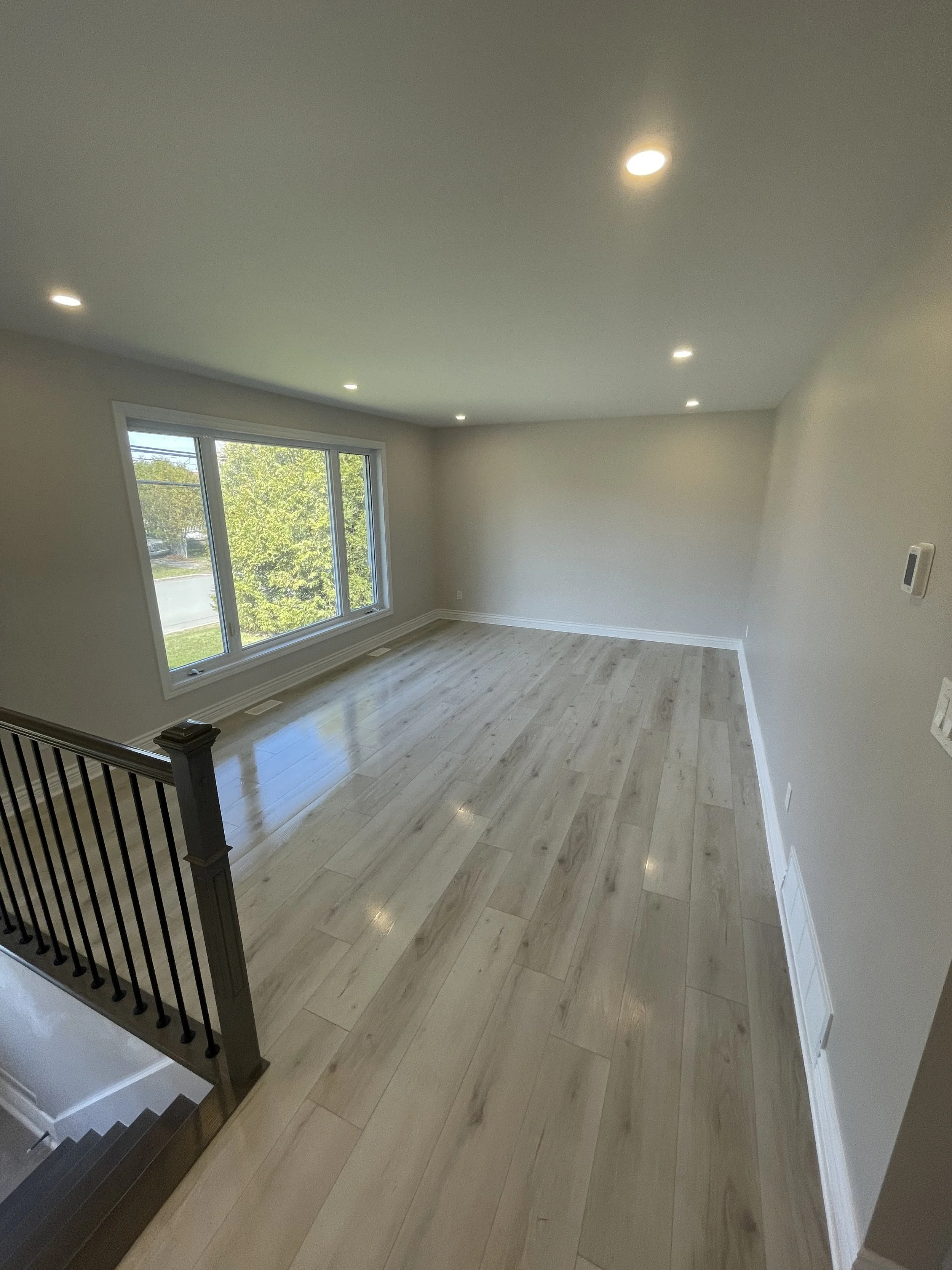 Empty living room with large window, hardwood floors, recessed lighting, and staircase with black railings.