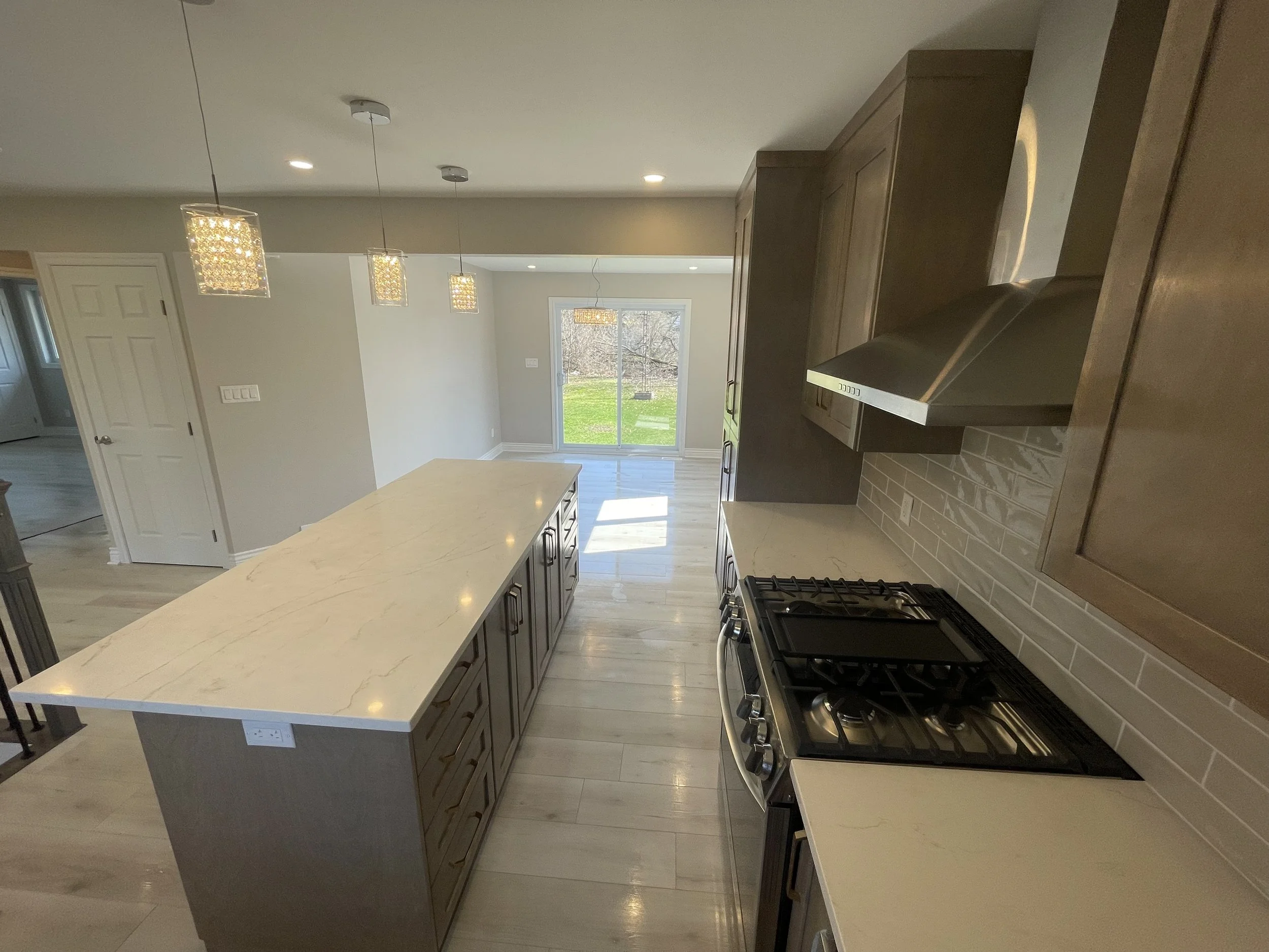 Modern kitchen with a large white marble island, wooden cabinets, stainless steel appliances, and pendant lights, open to a dining area with sliding glass door leading to a backyard.