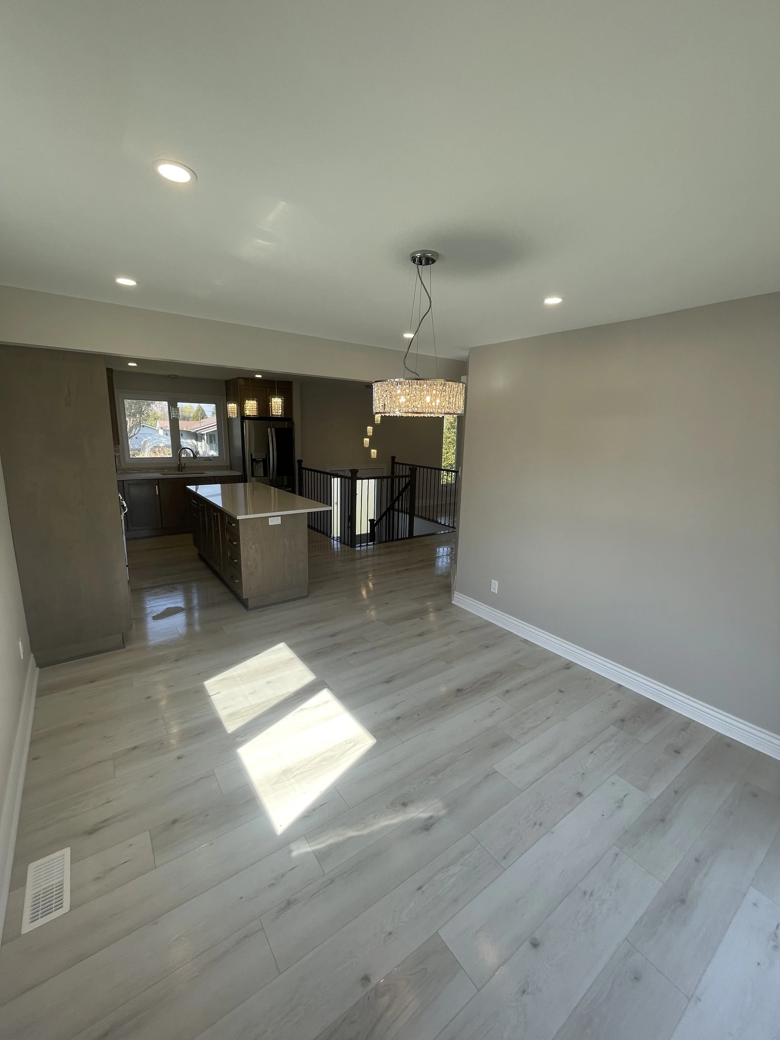 Empty living room with wooden floors, gray walls, recessed lighting, a window, and a staircase leading to the upper floor, with a chandelier hanging from the ceiling.