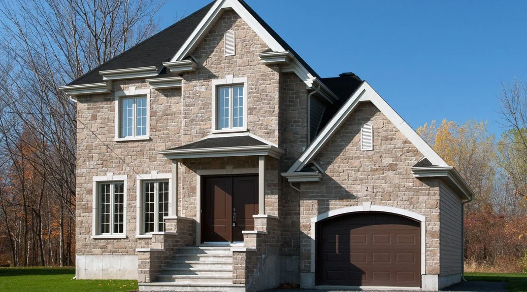 A two-story house with a stone facade, black roof, and white trim. It has a front porch with steps leading up to a dark brown door, multiple windows, and an attached garage with a dark brown door. The house is set against a backdrop of trees with fall foliage and a clear blue sky.