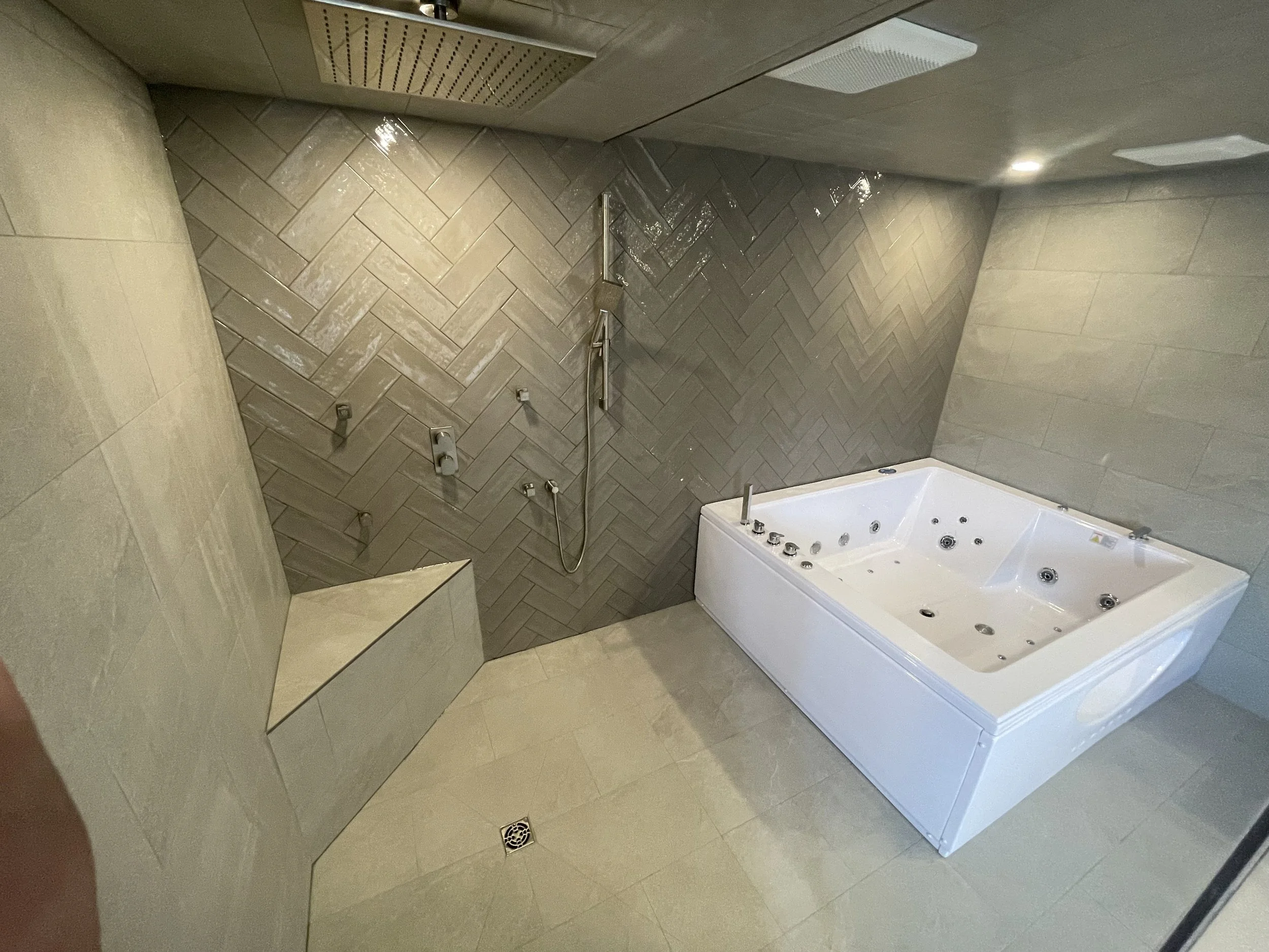 A modern bathroom with a jacuzzi tub featuring multiple jets, and a walk-in shower with a handheld showerhead, tiled walls, and a floor drain.