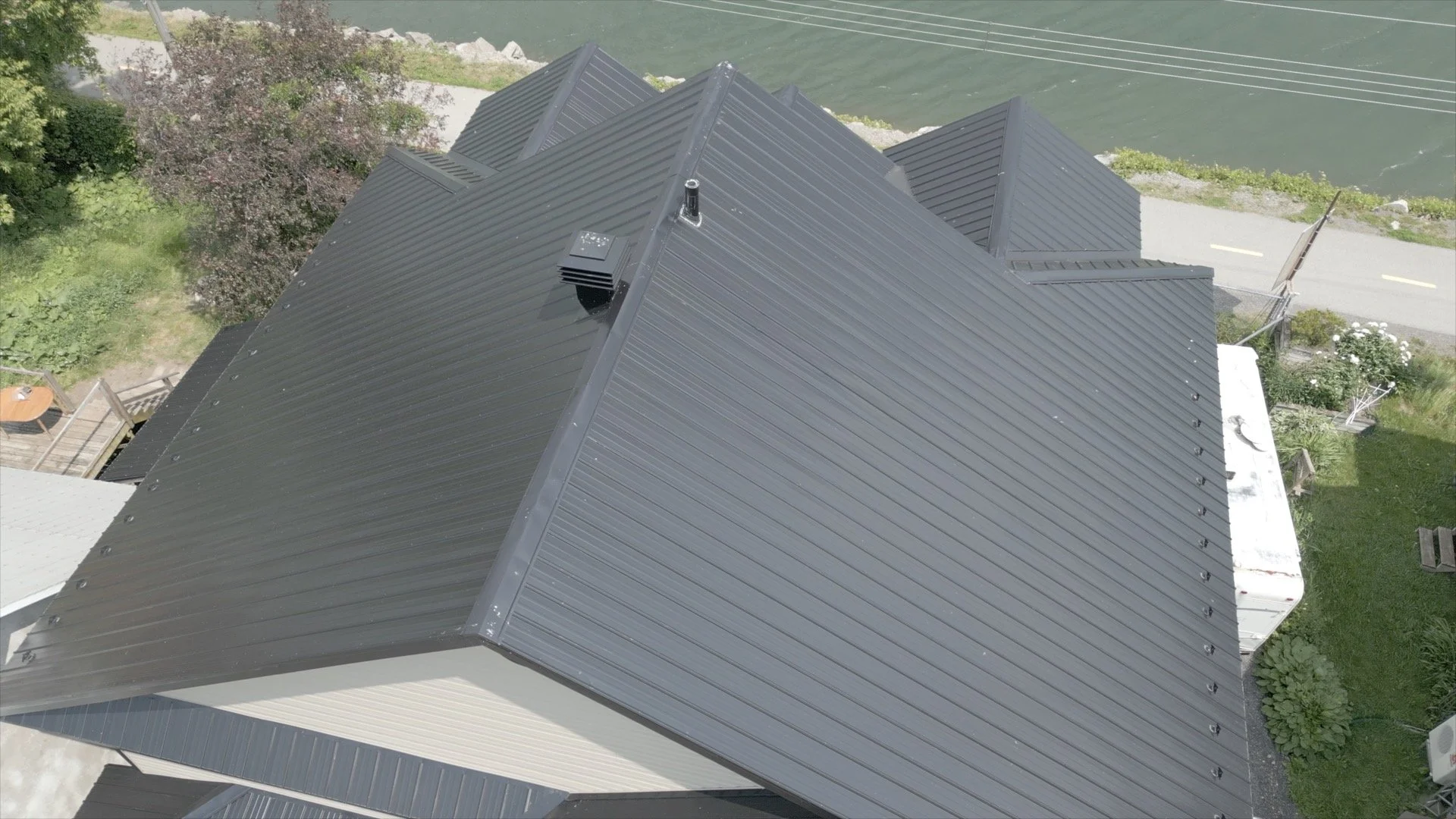 Top-down view of a house with a metal roof next to a body of water with trees and a road nearby.