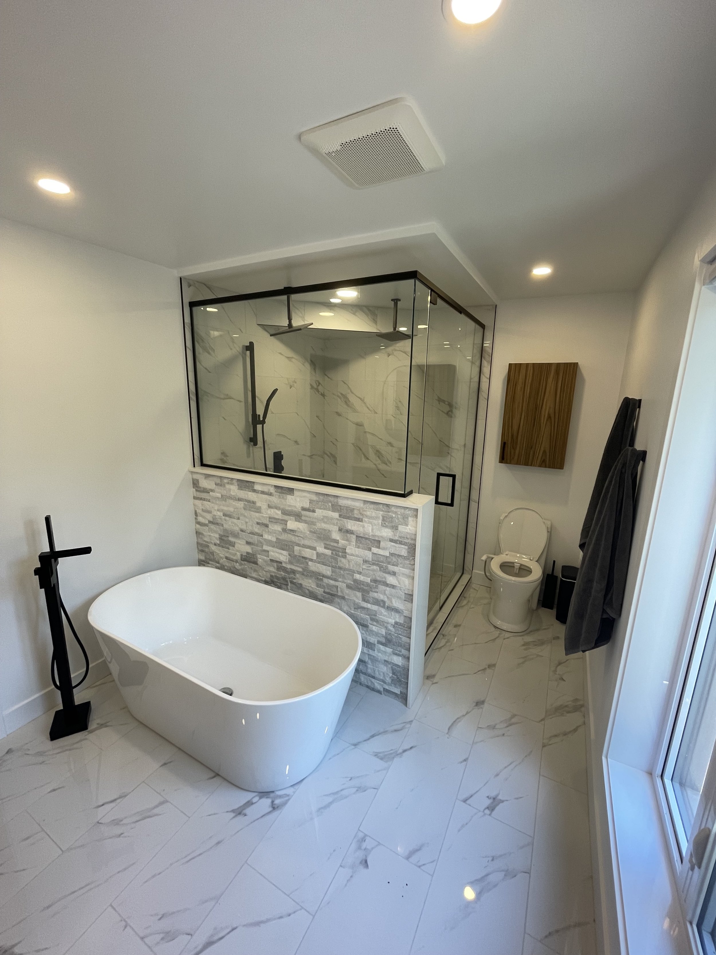 Modern bathroom with a free-standing bathtub, glass-enclosed shower with marble walls, a toilet, and a wooden cabinet. White marble-look floor tiles and recessed ceiling lights.