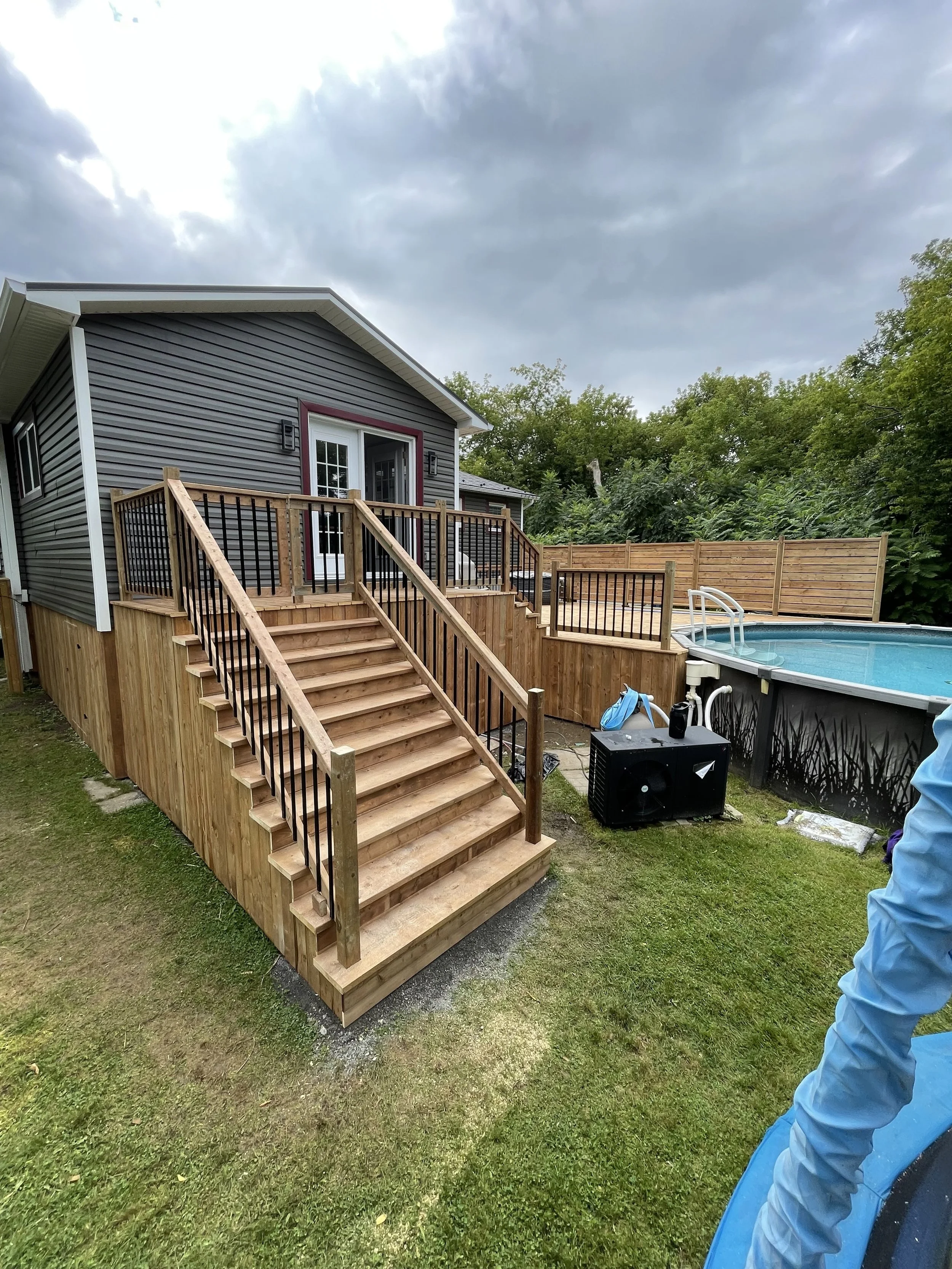 New wooden deck stairs leading to a house with gray siding and a sliding glass door, in a backyard with an above-ground pool and a fence, overcast sky.
