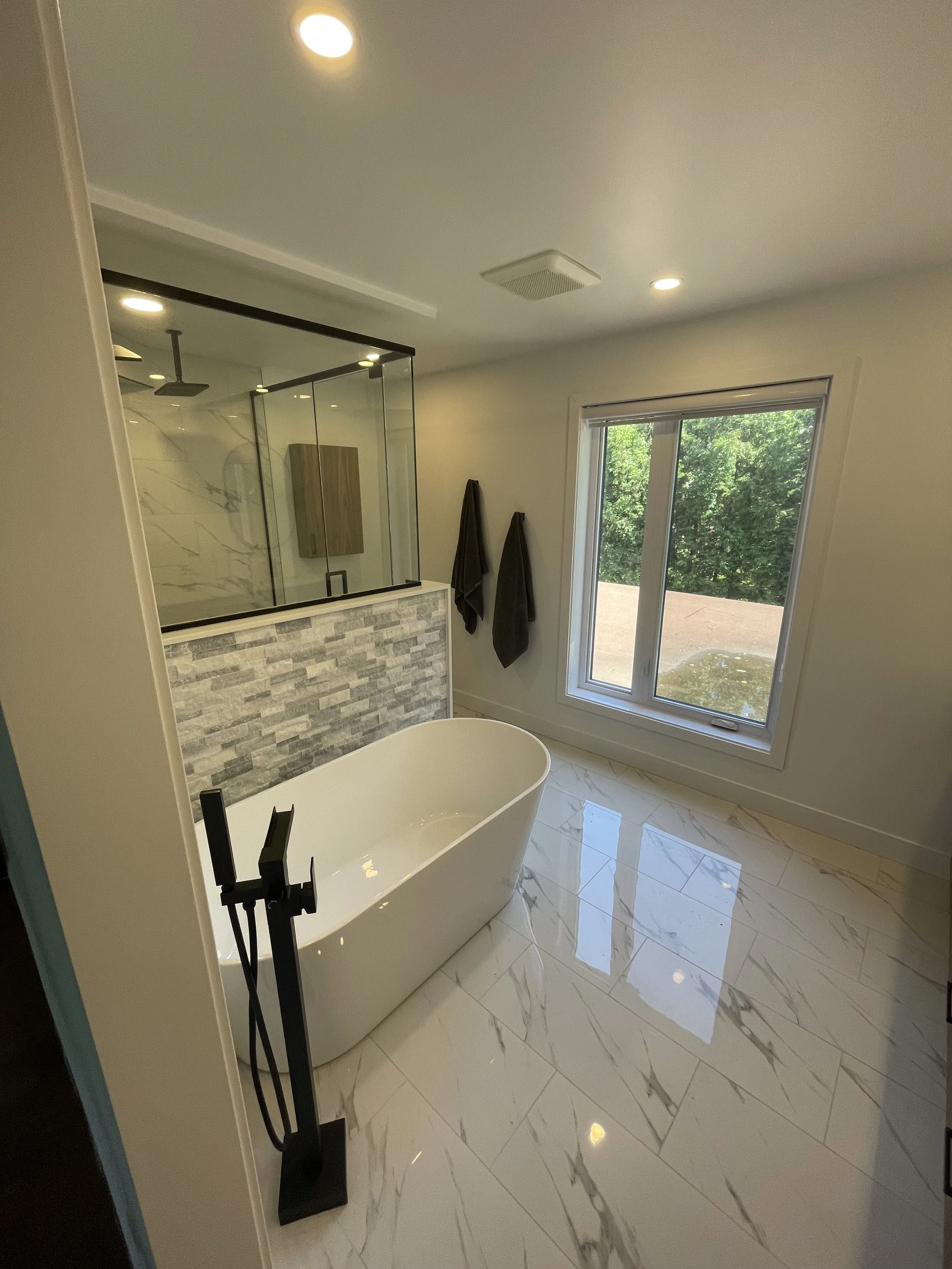 Modern bathroom with free-standing bathtub, large window, glass-enclosed shower, and marble tile flooring.