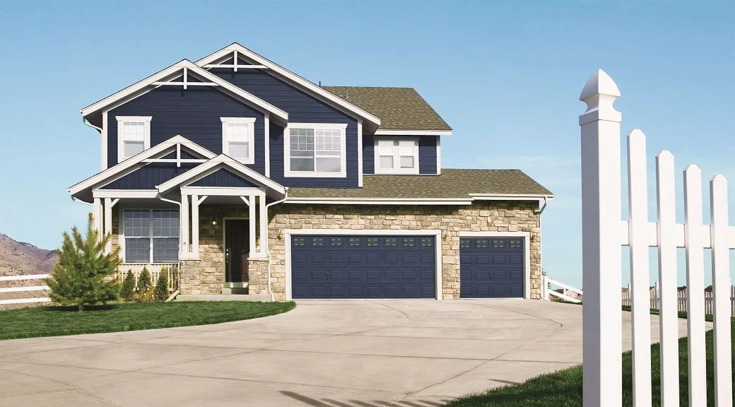 A two-story house with blue siding, white trim, a stone accent on the front, and an attached garage with three doors. There is a green lawn, small bushes, and a white fence on the right.