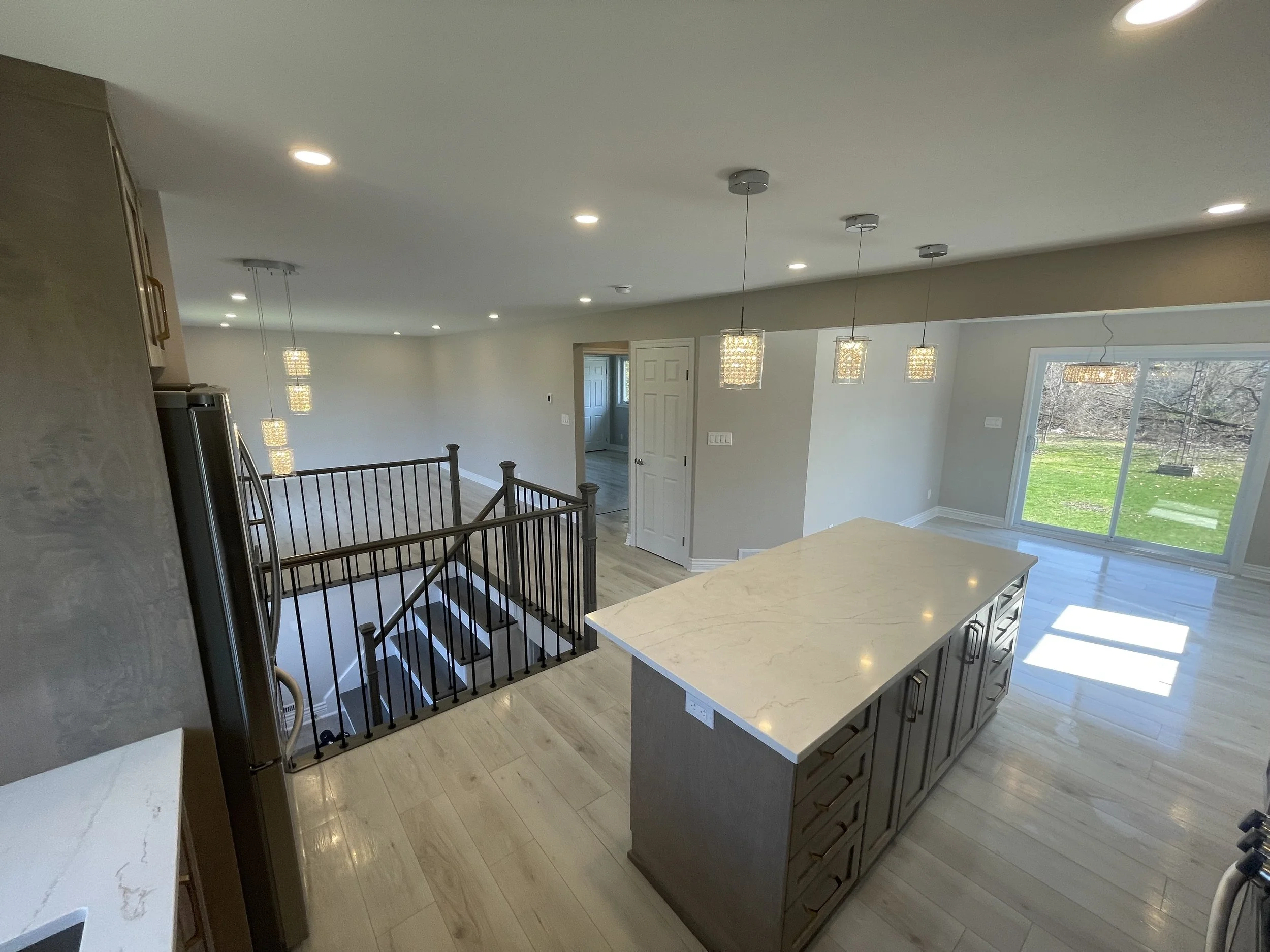 Empty modern kitchen with a large granite island, stainless steel refrigerator, and pendant lighting over the island. Sliding glass door leads to a backyard with grass and trees.