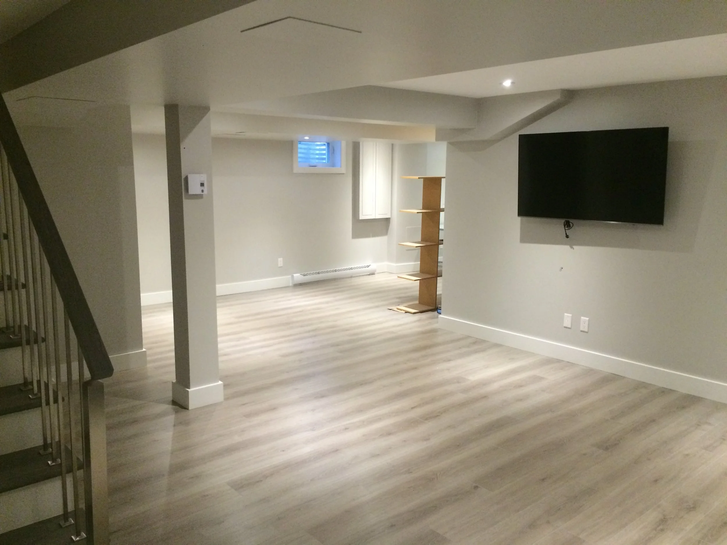 Empty basement with light-colored wood flooring, a mounted flat-screen TV, small window, staircase, and a wooden bookshelf.