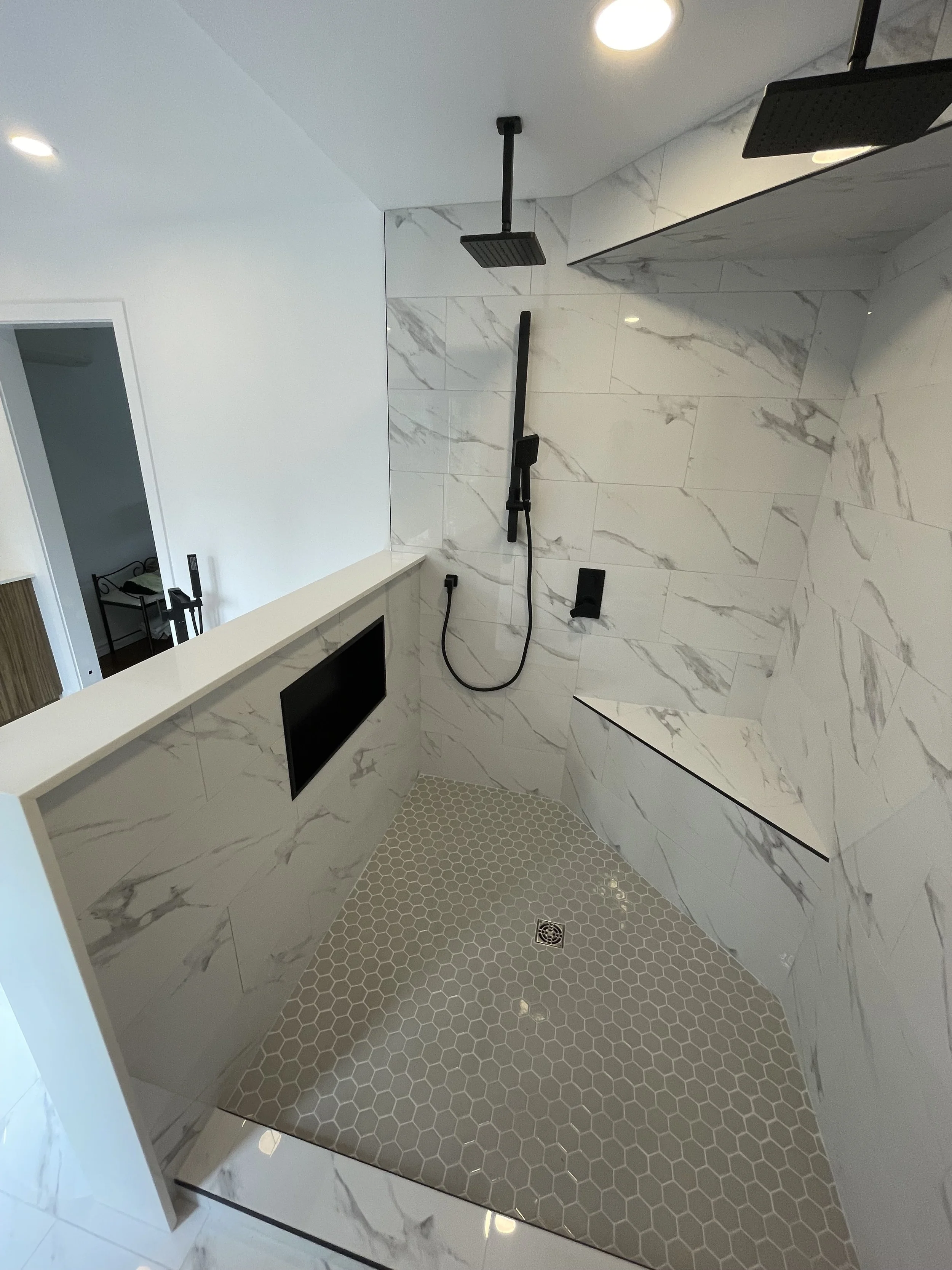 Modern walk-in shower with white marble tiles, two black showerheads, a handheld shower, a built-in niche, and a small black TV mounted on the wall.