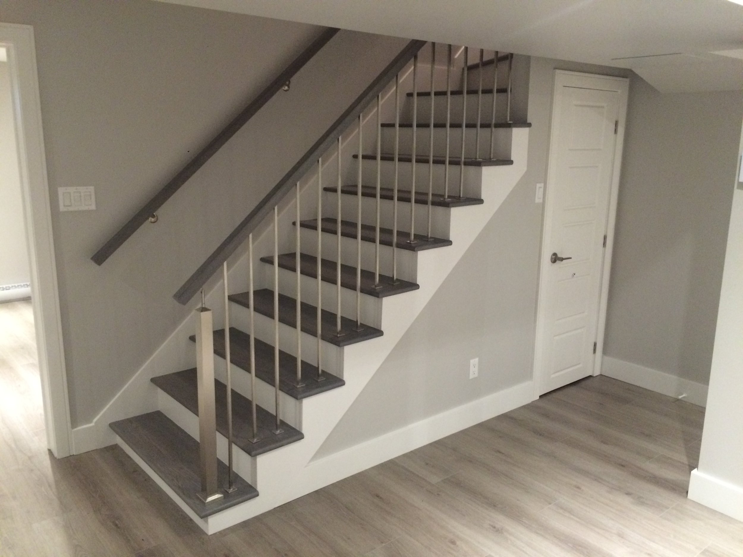 A modern staircase with dark wooden steps, gray handrails, and white spindle balusters in a house interior with light wood flooring and gray walls.