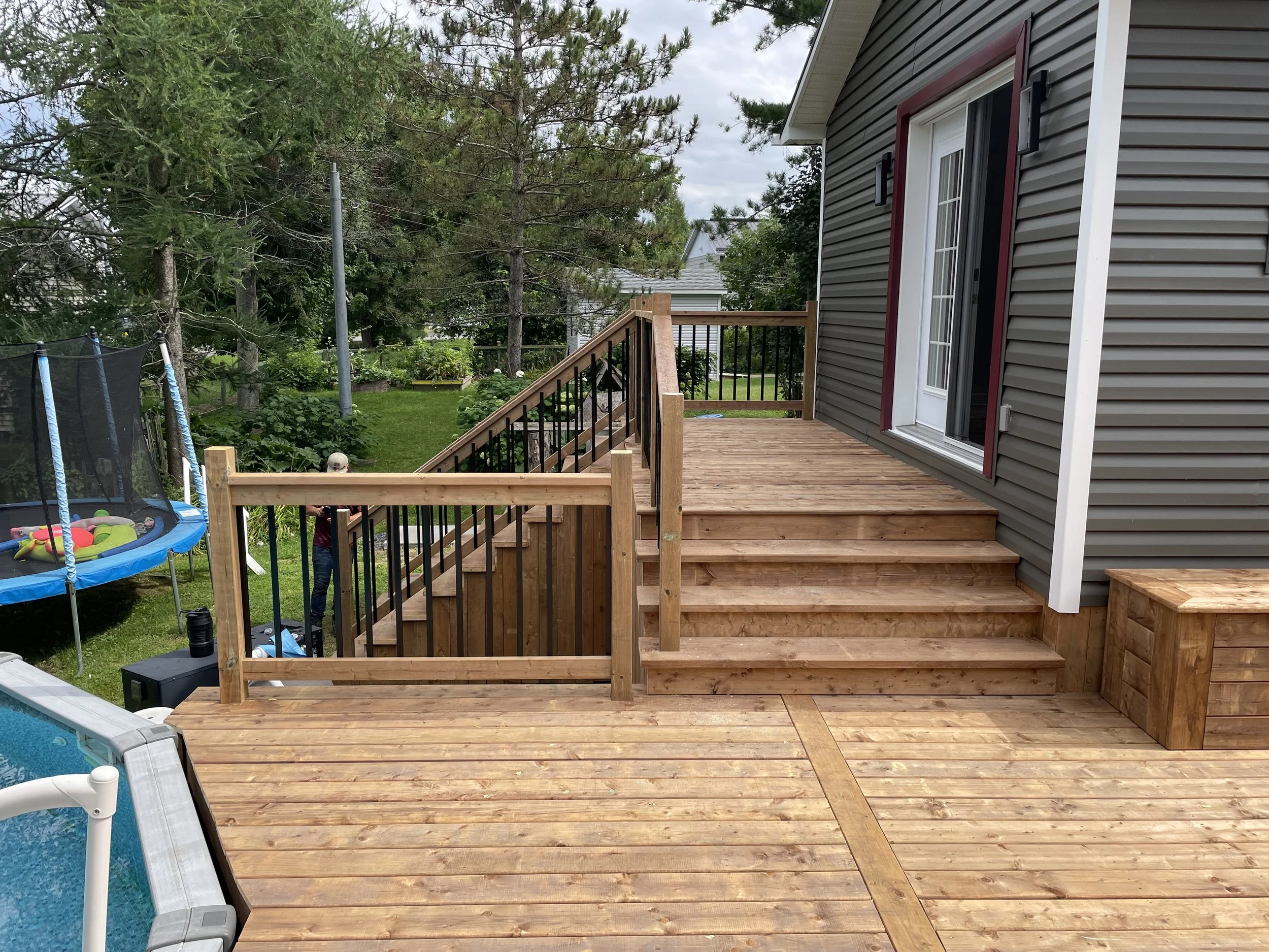 Backyard deck with stairs leading to grass yard, sliding glass door, and fenced area with children’s trampoline and trees.