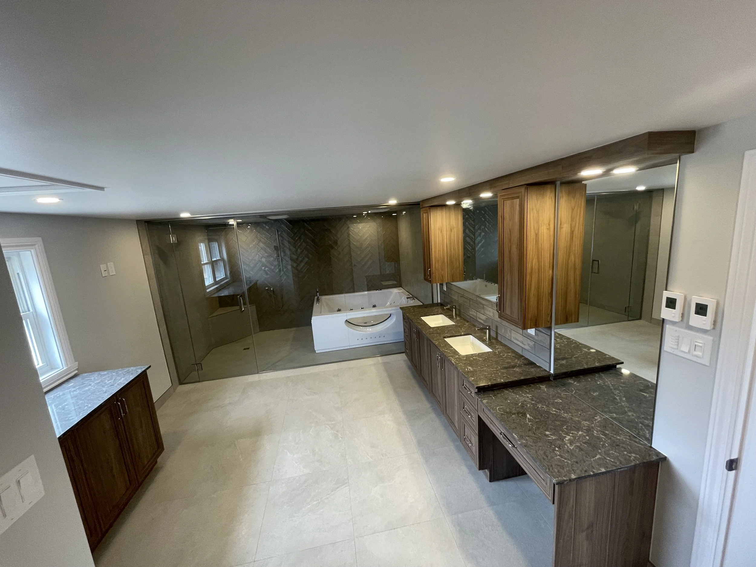 Modern bathroom with double vanity, dark granite countertops, wooden cabinets, a walk-in shower with glass doors, a soaking bathtub, and large beige tile flooring.