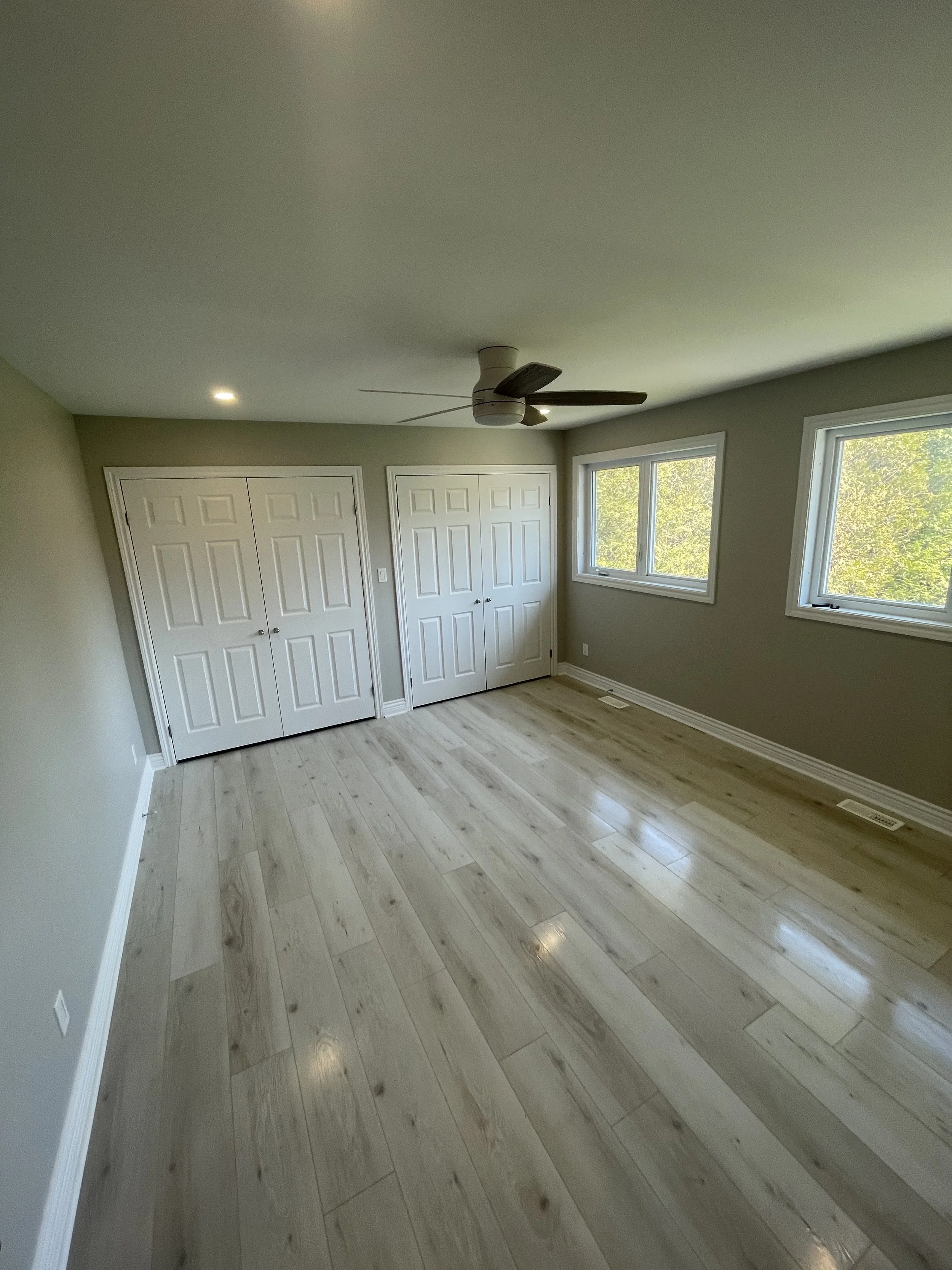 Empty bedroom with light wooden floors, green walls, two windows, two white closet doors, ceiling fan, and recessed lighting.