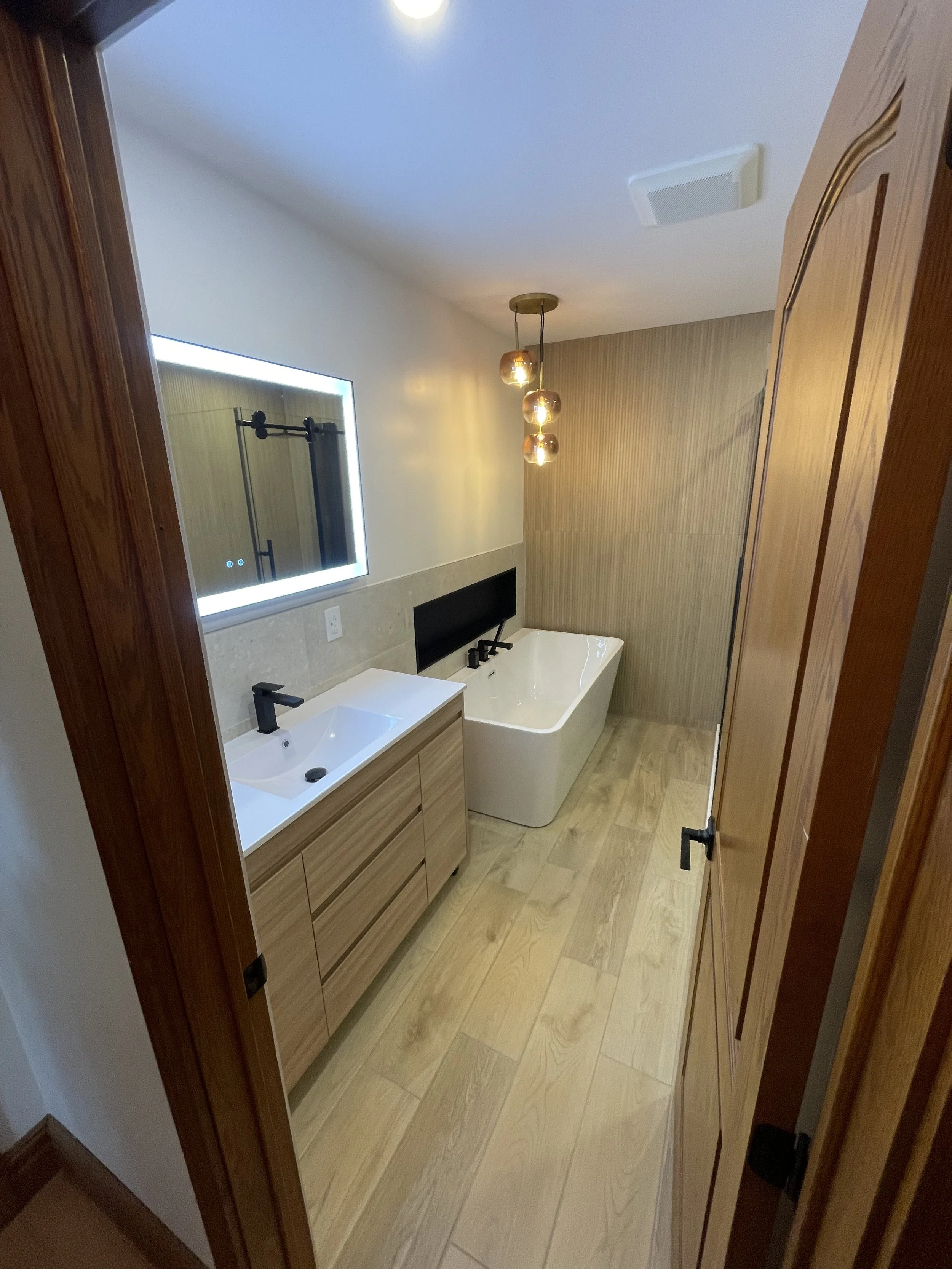 Modern bathroom with wooden vanity with black fixtures, a freestanding bathtub, a large illuminated mirror, beige tiled walls, wooden flooring, and a hanging copper pendant light fixture.