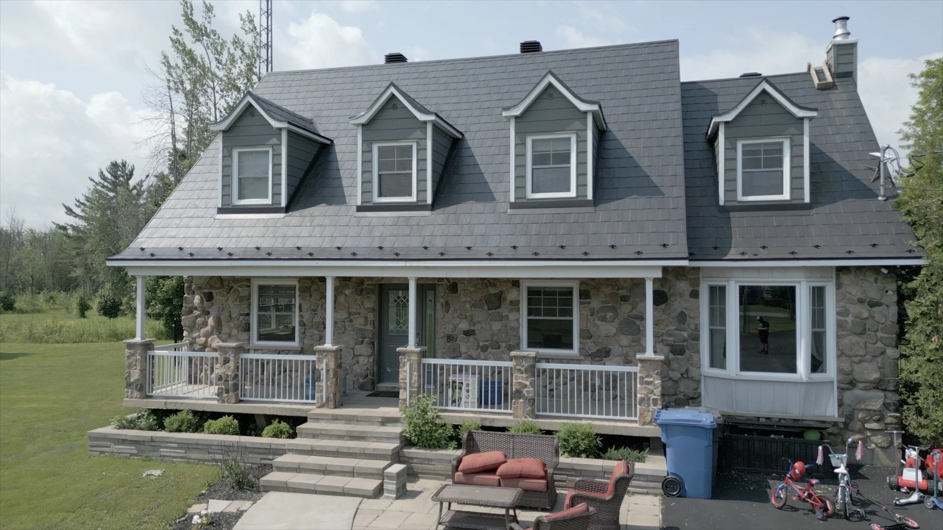 Front view of a two-story house with stone exterior walls, gray roof with three dormer windows, front porch with white railing, outdoor furniture, and children's bikes in driveway.