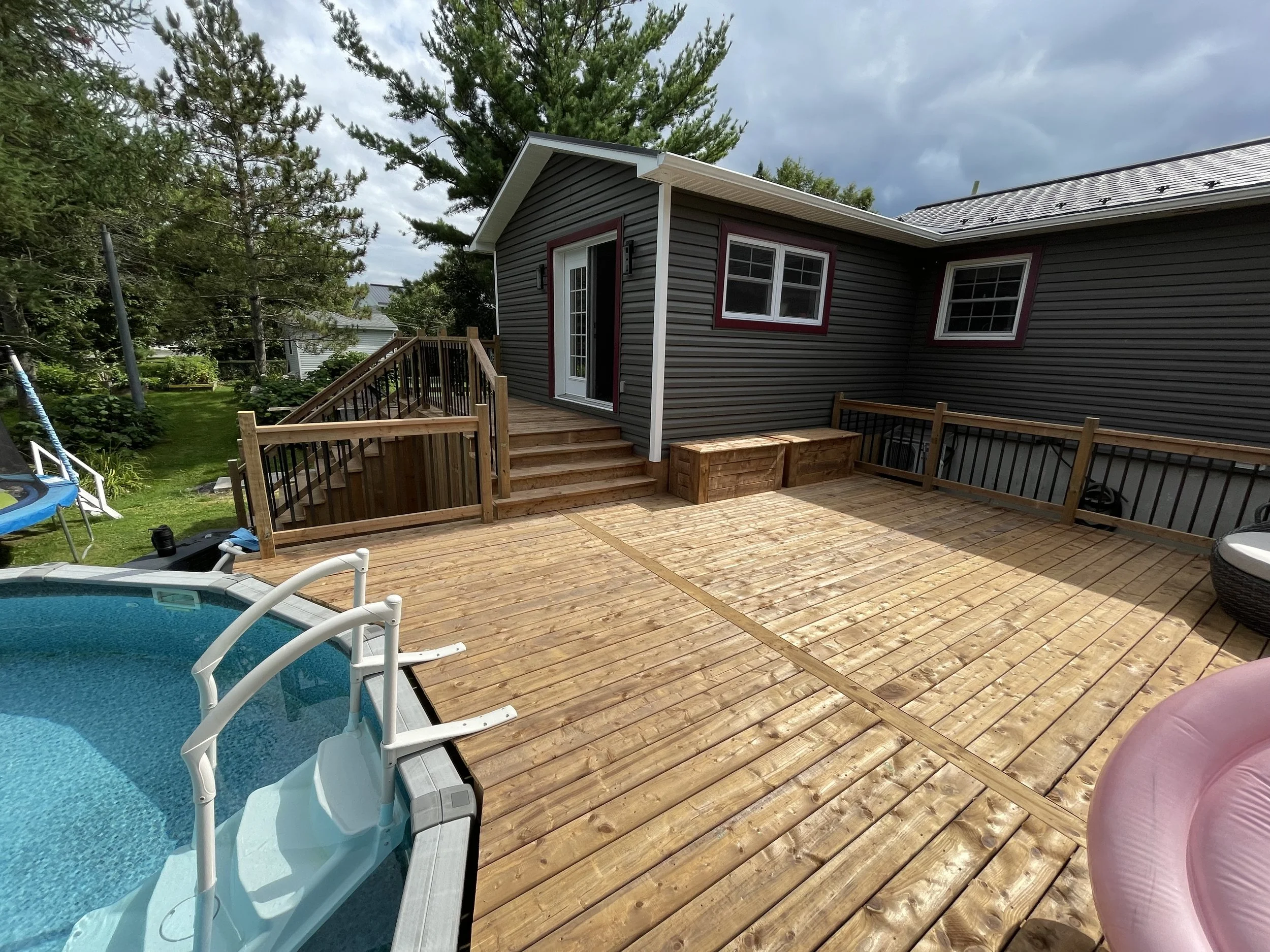 Backyard deck with a small pool, wooden stairs, and a house with dark gray siding and white trim, surrounded by trees under a partly cloudy sky.
