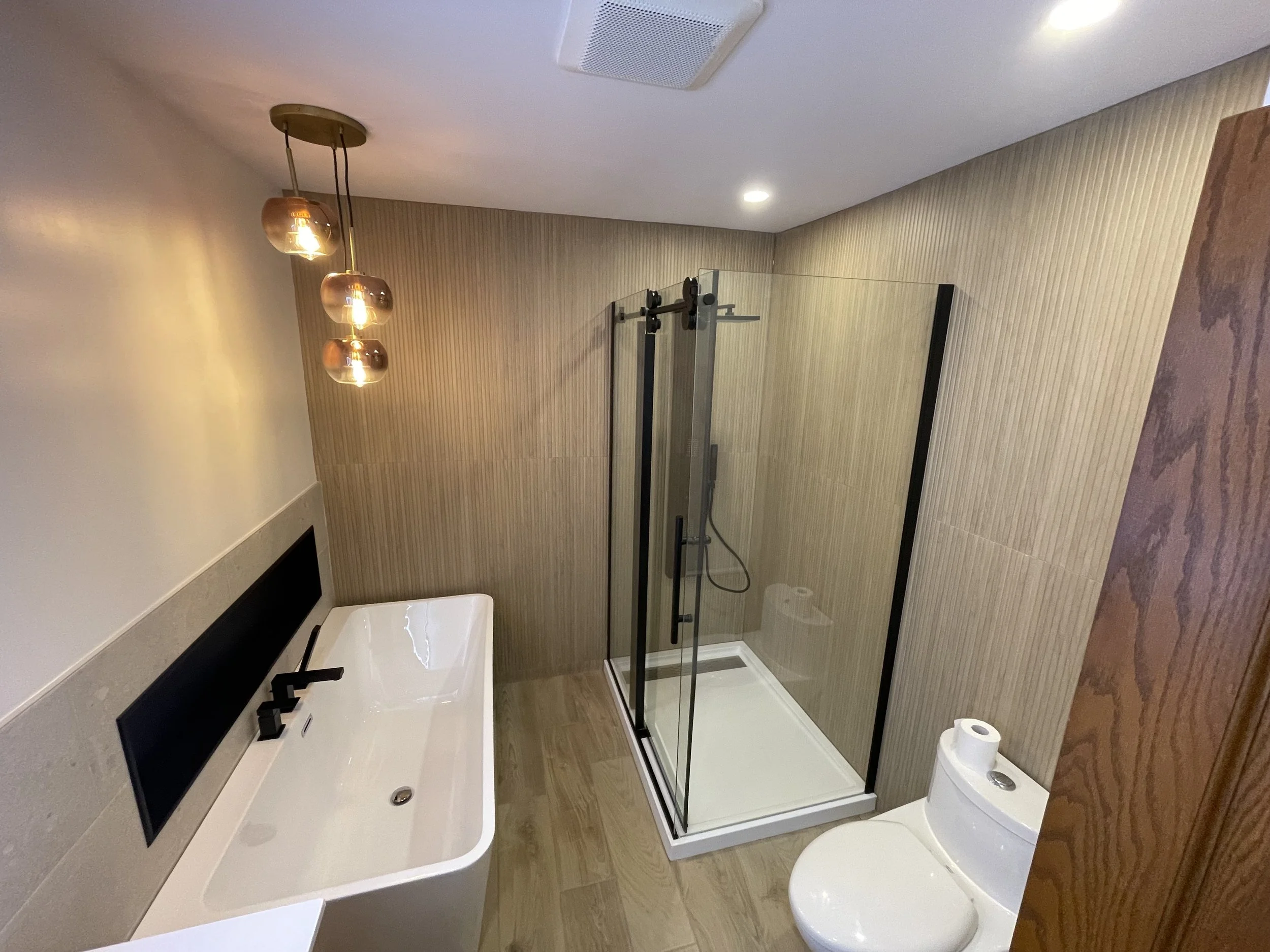 Modern bathroom with walk-in shower, white bathtub, toilet, beige wall tiles, wooden elements, and hanging pendant lights.