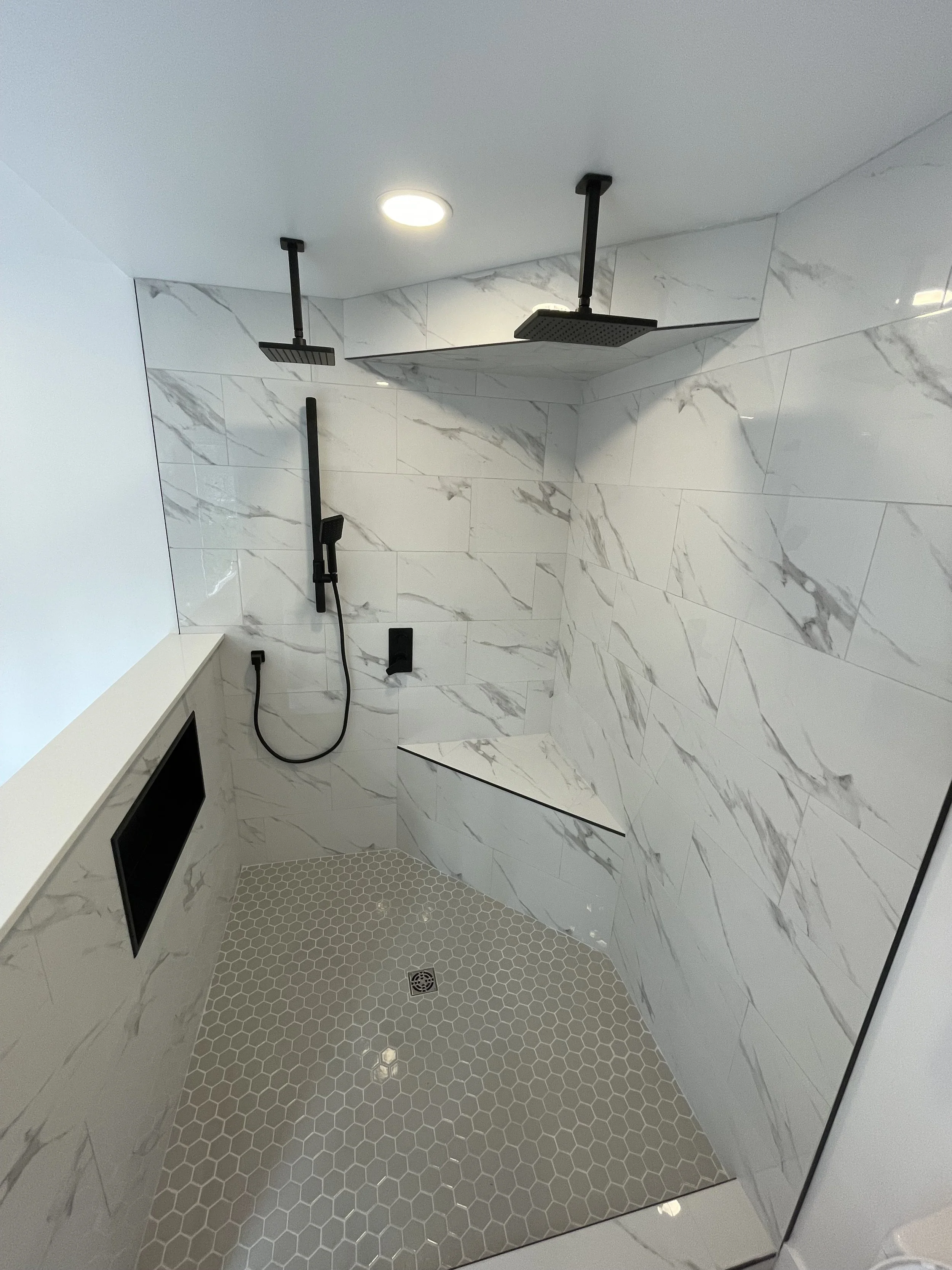 Modern walk-in shower with marble walls, two rainfall showerheads, a handheld shower, a built-in bench, and a floor drain with small hexagonal tiles.