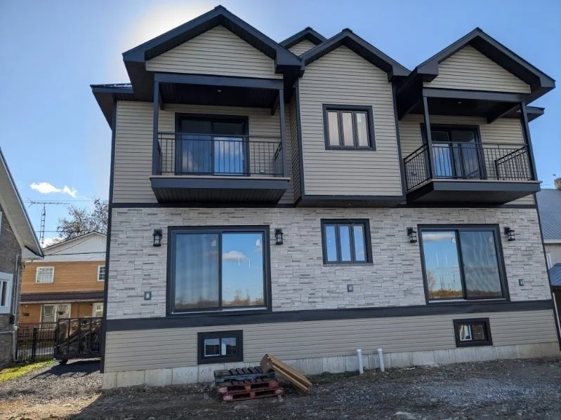 Three-story modern house with beige siding, stone accents, and small balconies on the upper floor, set against a blue sky.