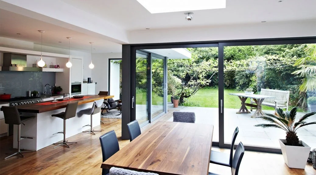 Modern kitchen and dining area with large sliding glass doors leading to a lush garden outdoor patio with table, bench, and potted plants.