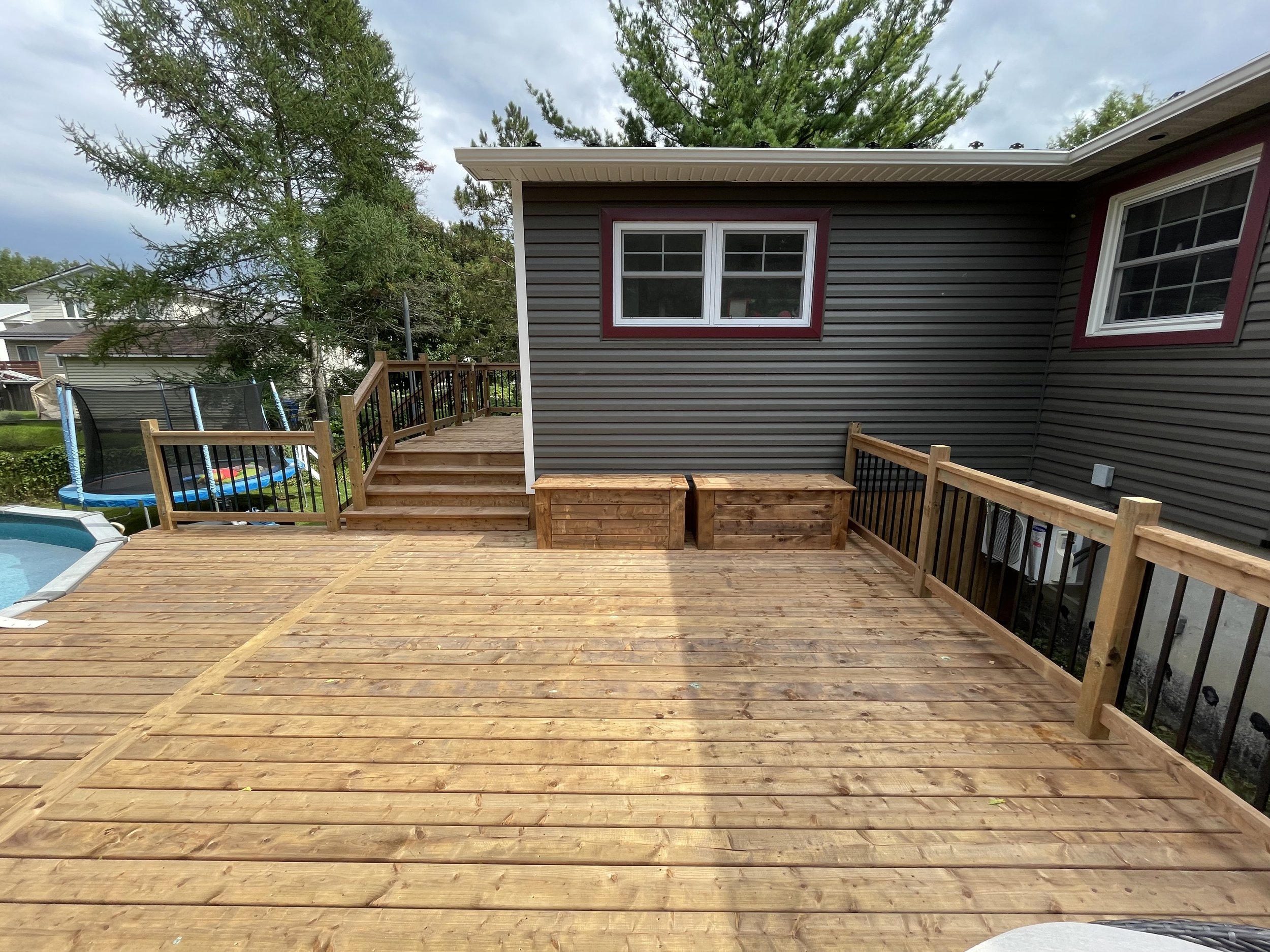 Wooden deck with steps leading up to another deck area, black railing, two wooden planters, and a gray house with red window frames in the backyard. Trampoline and pool are partially visible in the yard.