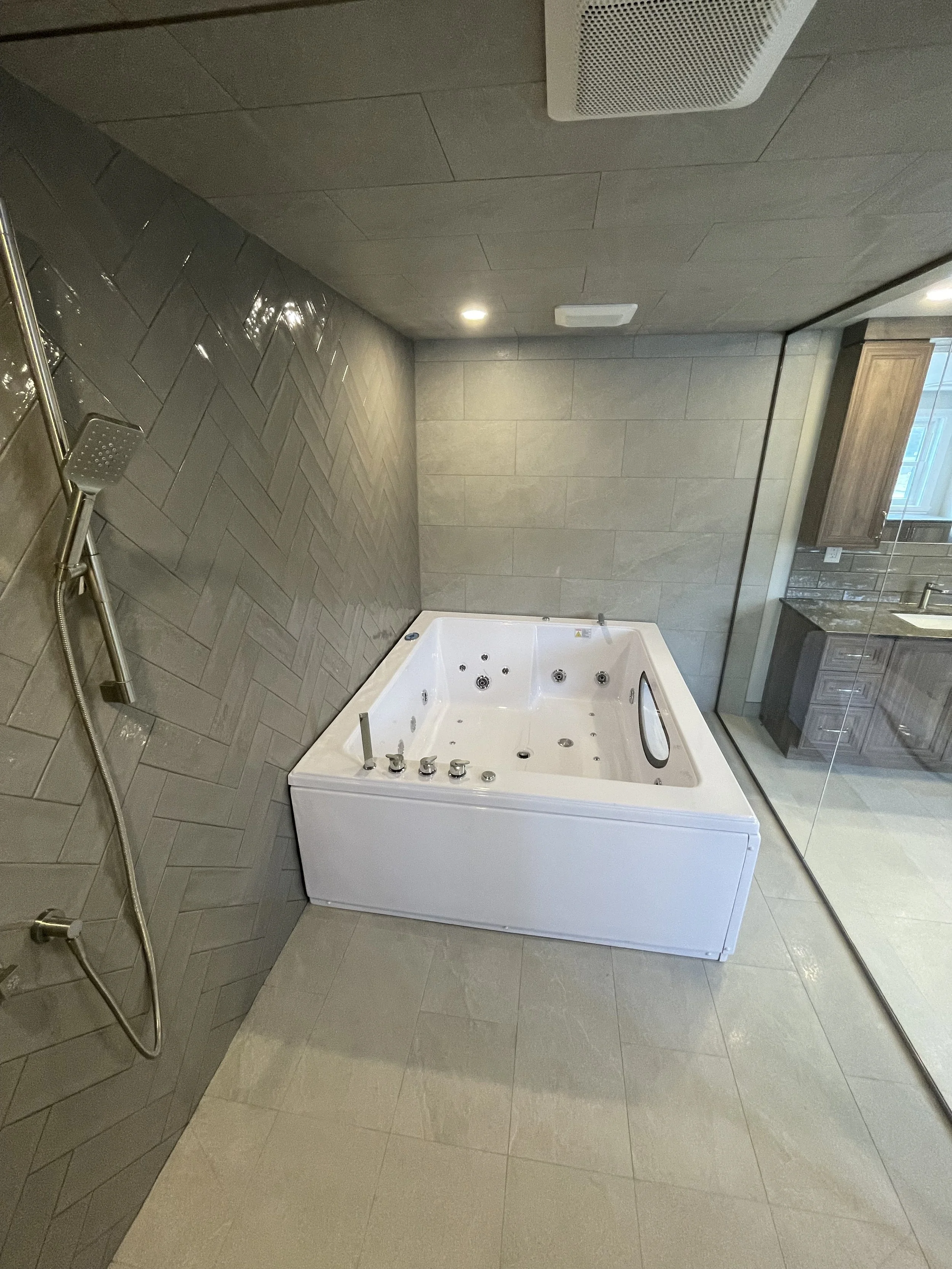 A bathroom with a white hot tub, gray tiled walls, and a shower head on the left wall. The bathroom has a beige tiled floor and a glass partition separating it from a vanity area with a wooden cabinet and a mirror.