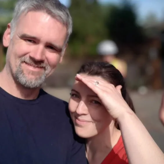 man with grey hair smiles towards camera while brown haired beautiful woman shields her eyes from the sun and smiles
