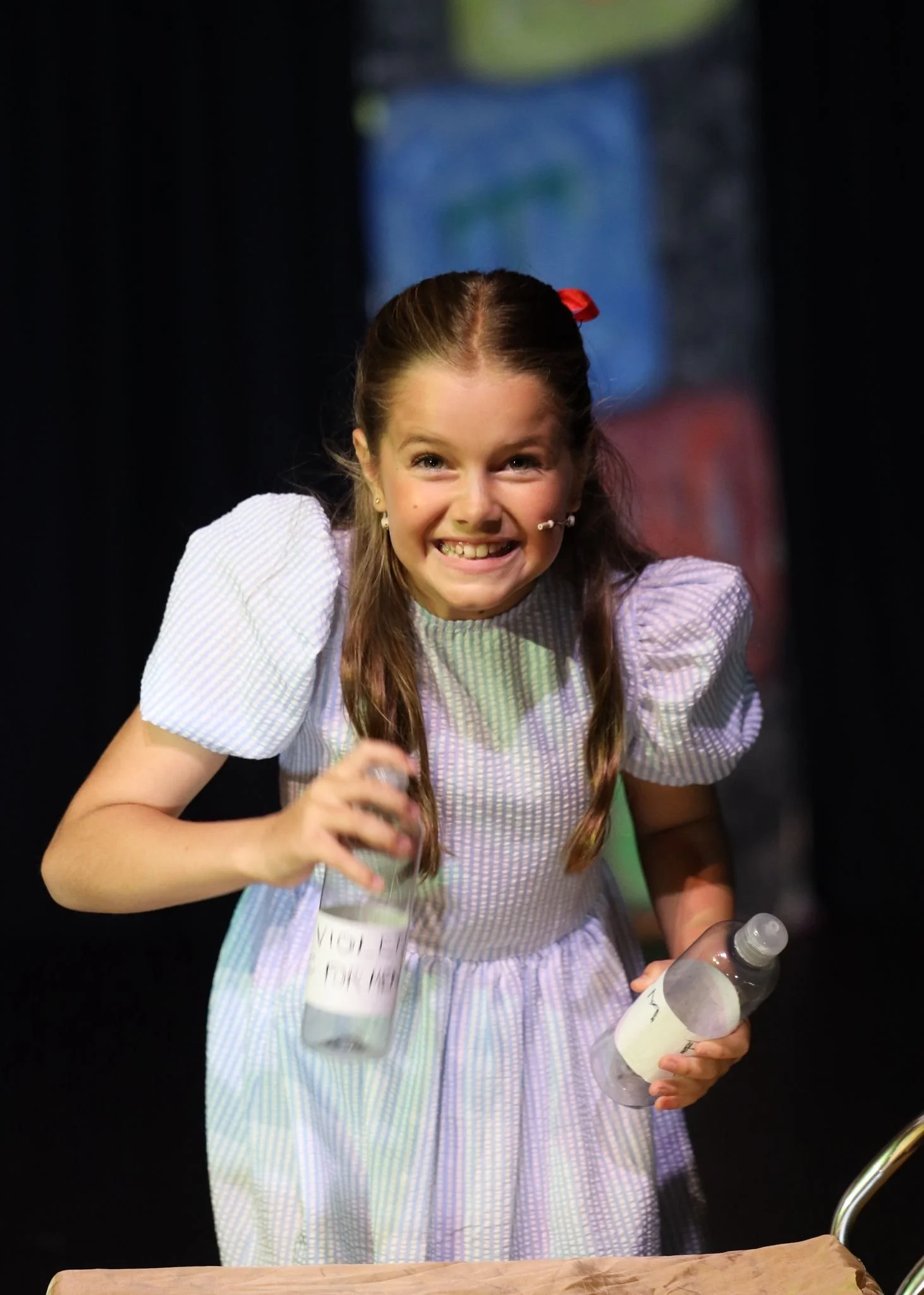 Young girl with pigtails and a big smile, holding clear water bottles with handwritten labels, on stage with a dark background.