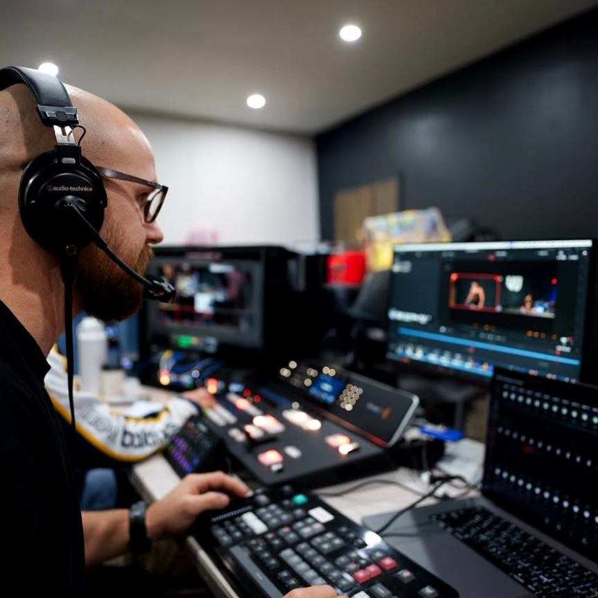 A person wearing headphones and glasses operating a video editing or streaming setup with multiple monitors and control panels.