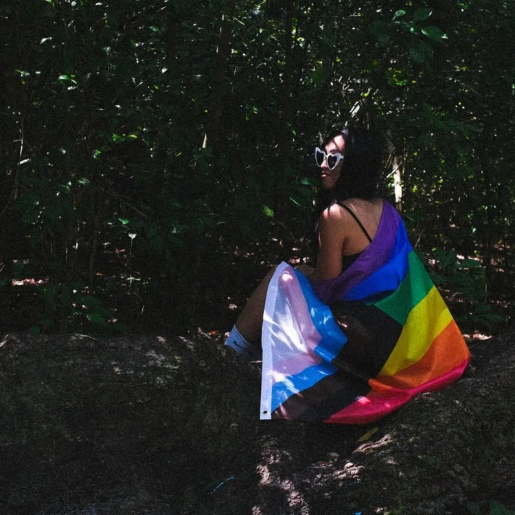 Person sitting on a rock in a forest, wearing sunglasses and a rainbow pride flag as a dress.