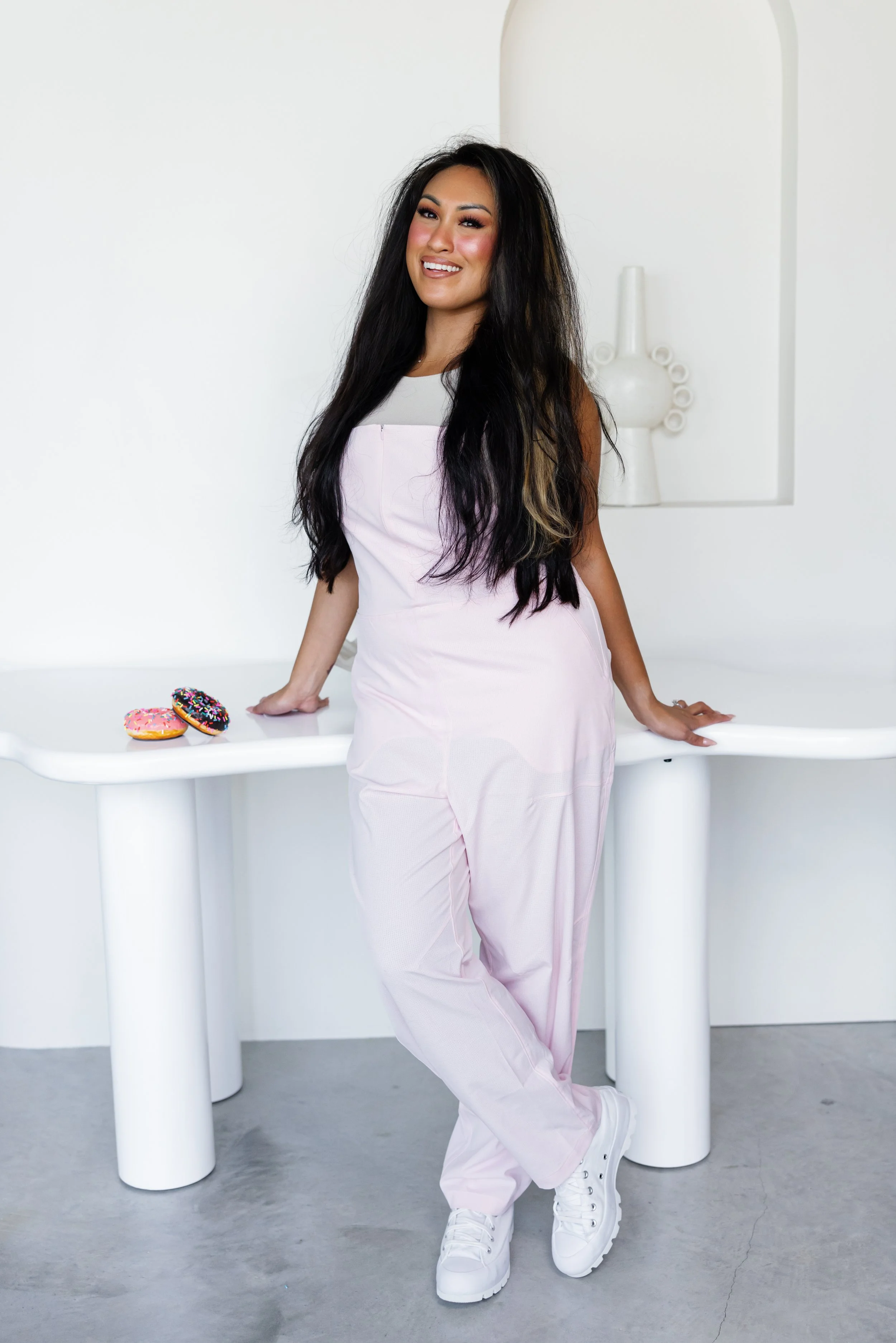 A woman with long dark hair and a light pink outfit standing next to a white table with two decorated donuts on it, in a minimalist white room.