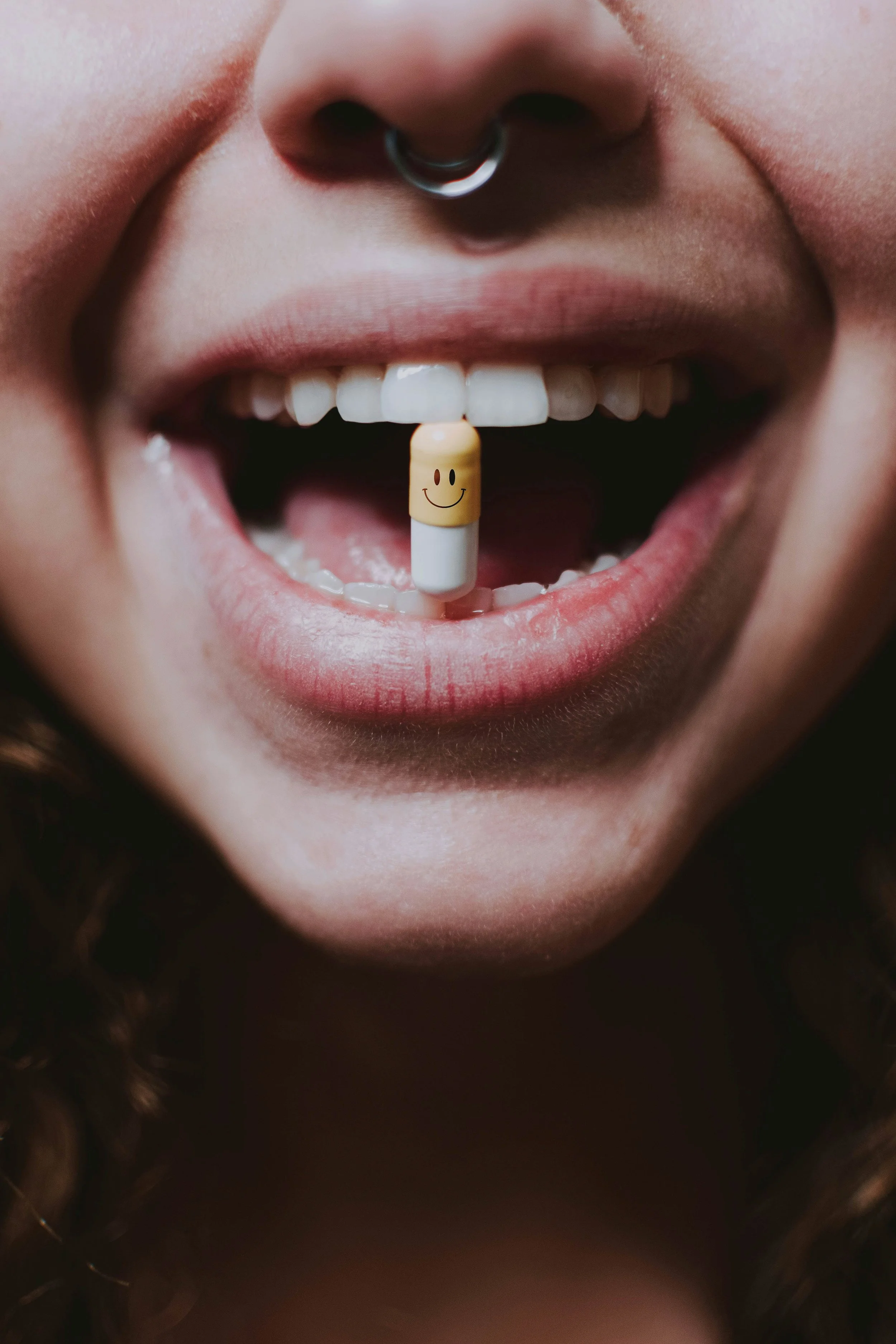 Close-up of a person’s open mouth with a yellow capsule pill with a smiley face design balancing on their lower teeth.