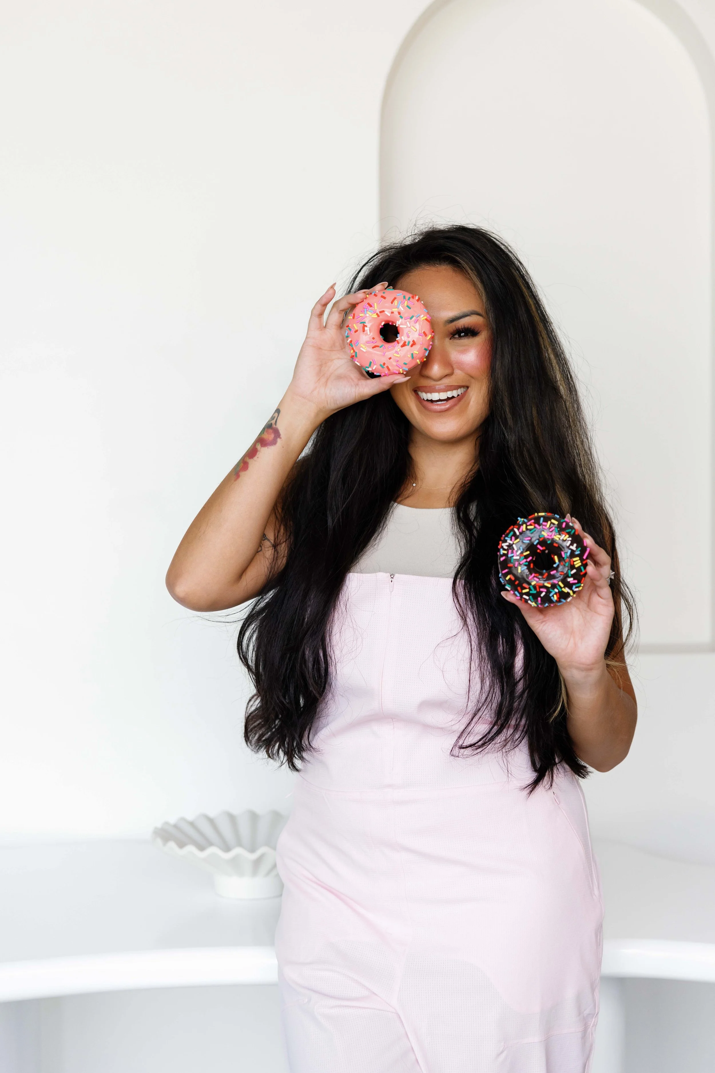 A woman with long dark hair smiling and holding two donuts with pink and chocolate icing and colorful sprinkles, one covering her right eye.
